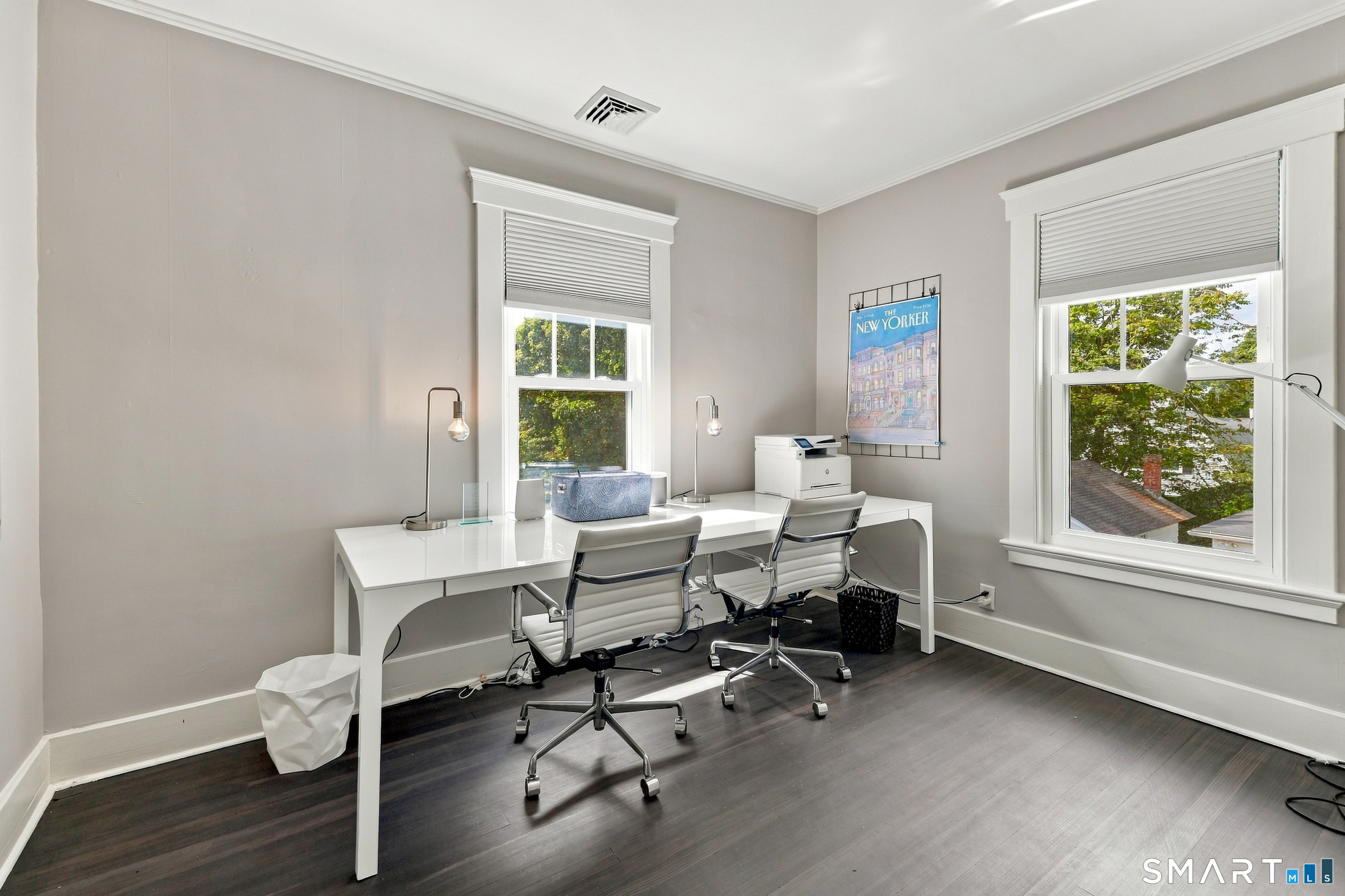 Furnished home office with a long white desk, two rolling chairs, and large windows.