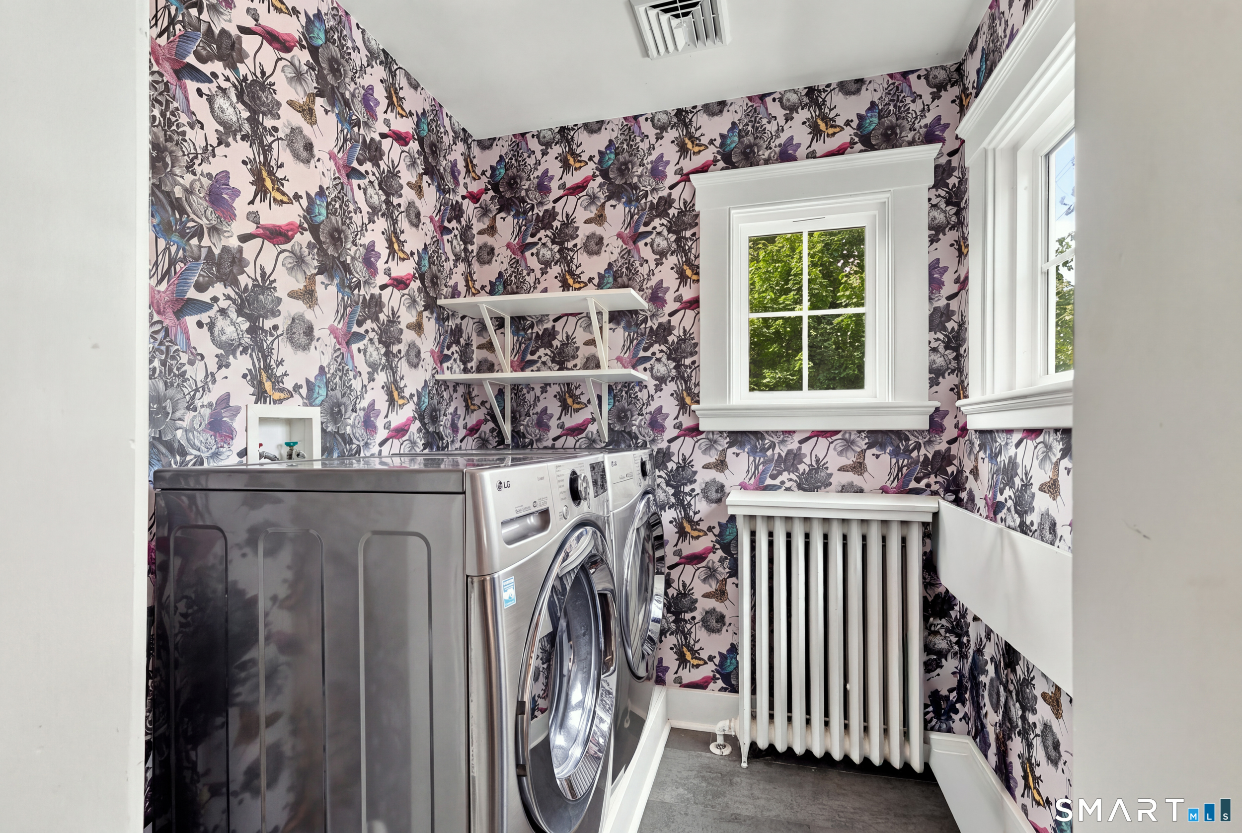Laundry room with front-loading washer and dryer, colorful bird-and-floral wallpaper, windows, and a radiator.