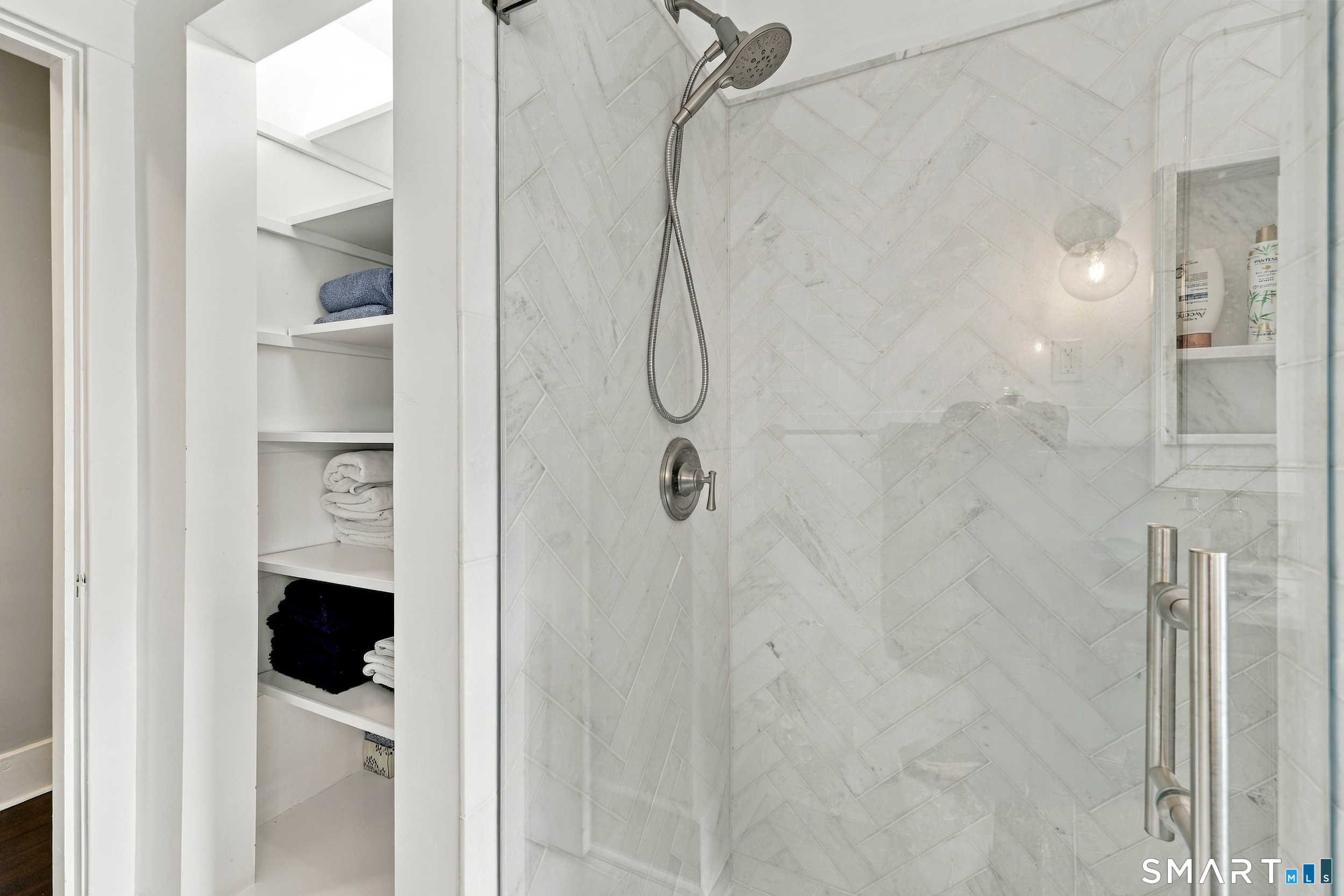 Glass-enclosed marble shower beside open shelves stocked with towels.