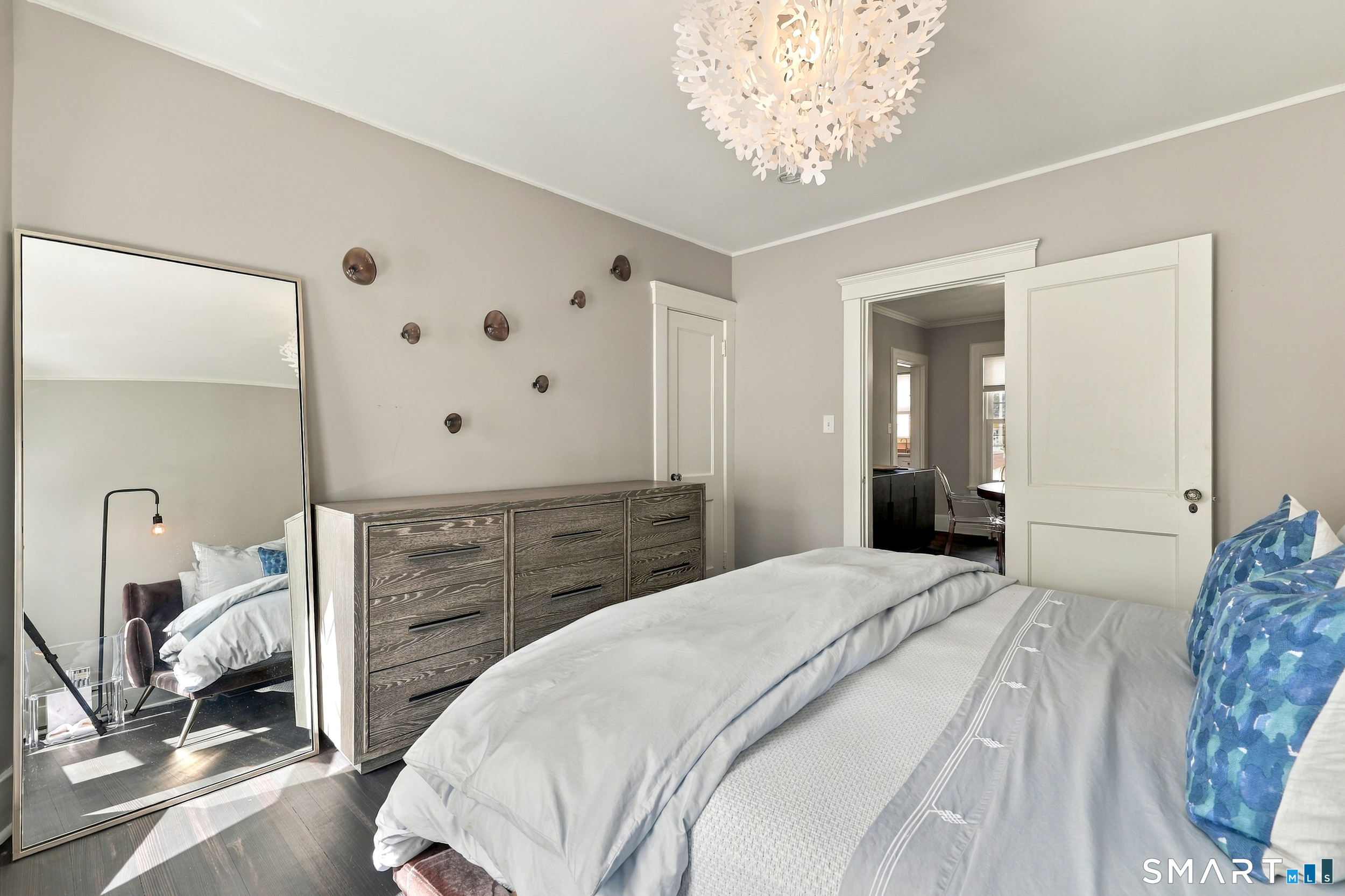 Bedroom with gray bedding, blue accent pillow, dark wood dresser, and sculptural chandelier.