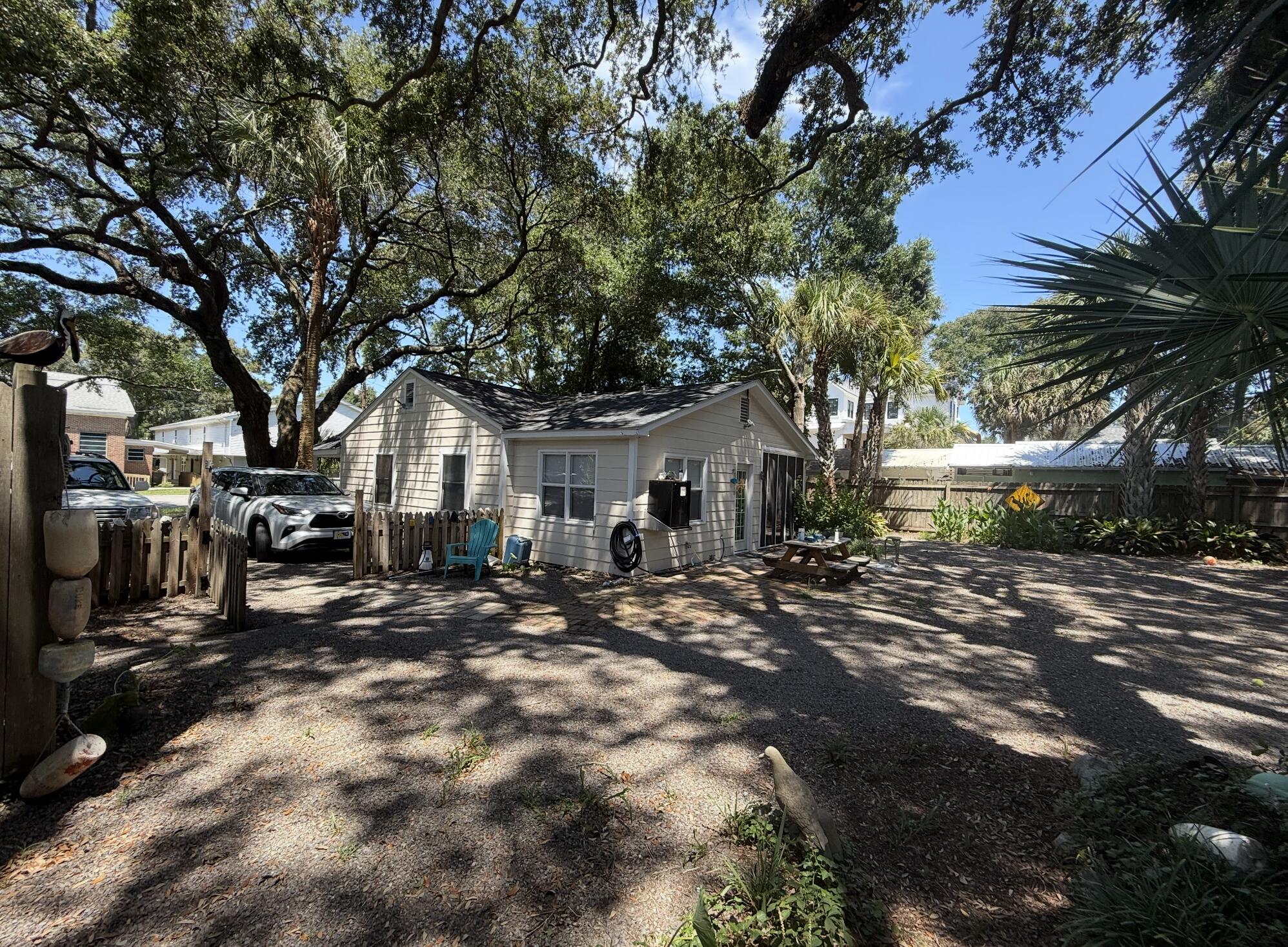 Small light-colored house beneath mature oak trees, with a gravel yard and picket fence.