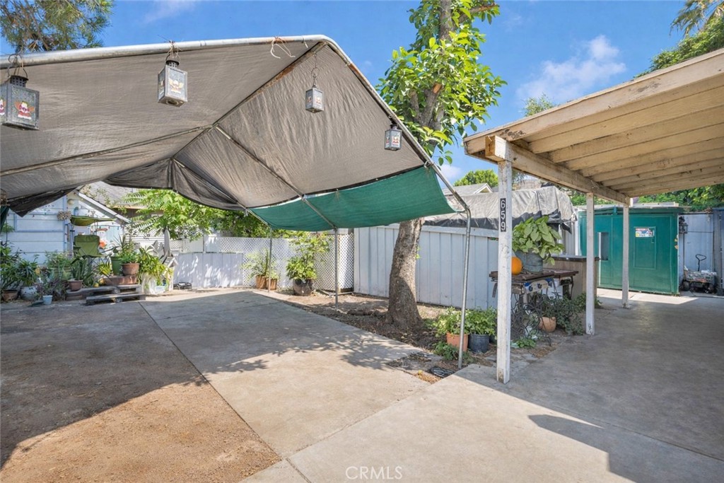 Paved outdoor area beneath fabric shade canopies, with a mature tree and green storage shed.