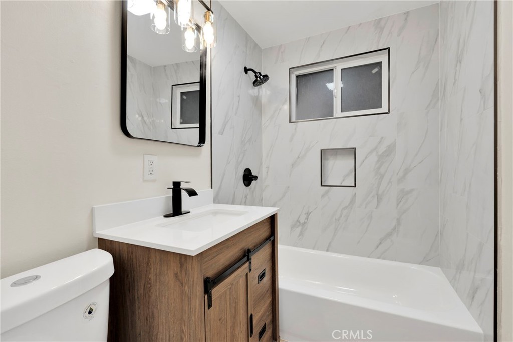 Bathroom with a wood vanity, black faucet, marble-look tub surround, high window and recessed niche.