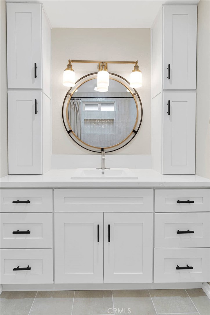 Bathroom vanity with a round mirror, brass lighting, and white cabinetry with black pulls.