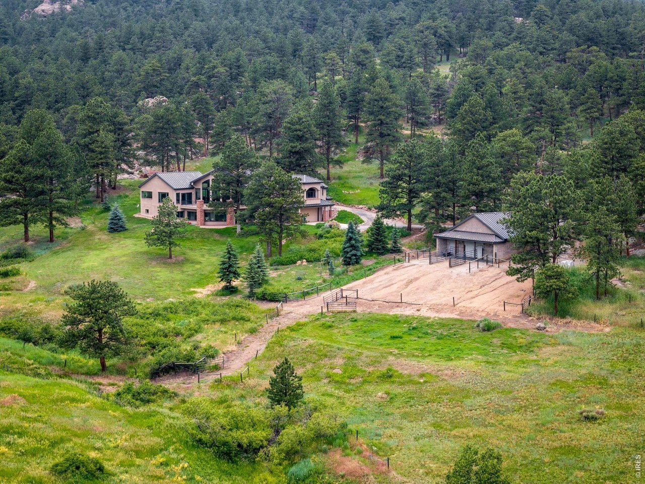 Aerial view of a mountain home surrounded by pine forest, with a detached outbuilding and dirt drive.