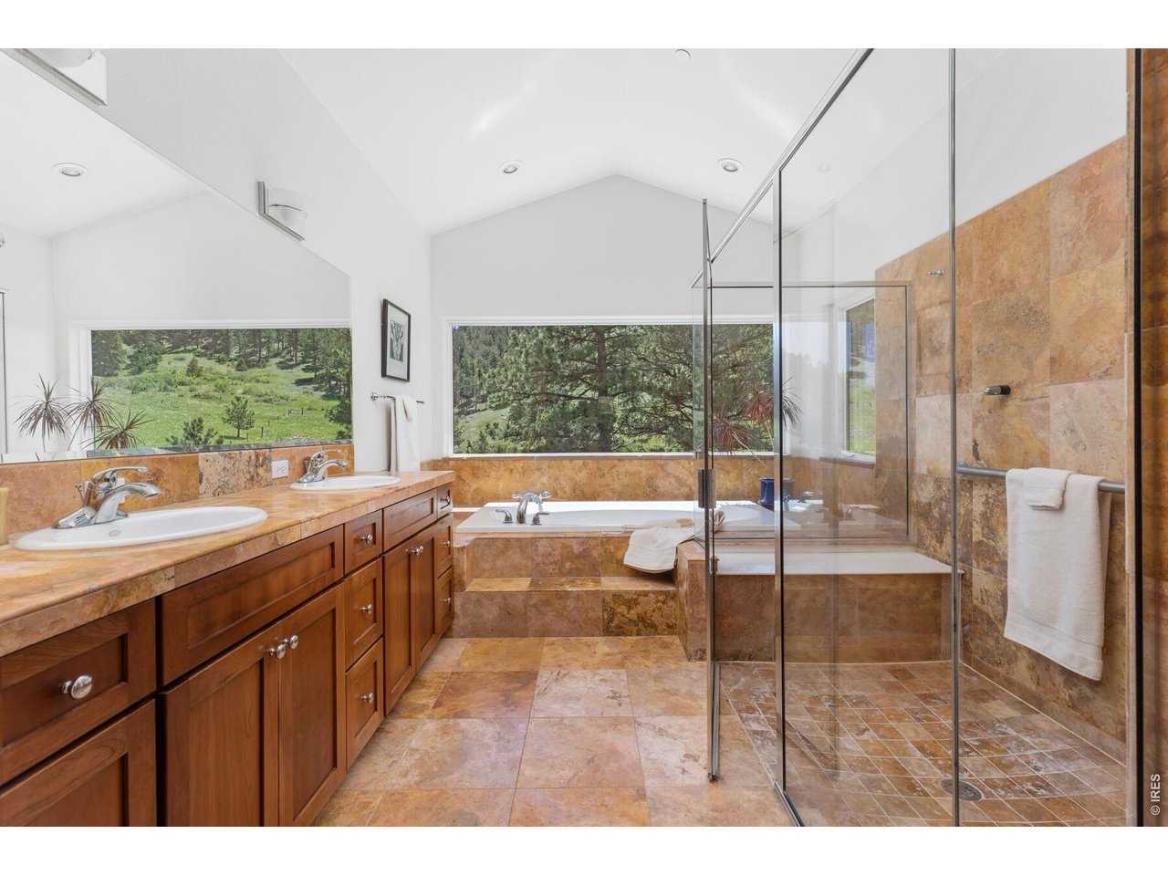Bathroom with double vanity, stone tile, soaking tub, and glass shower overlooking a green meadow.