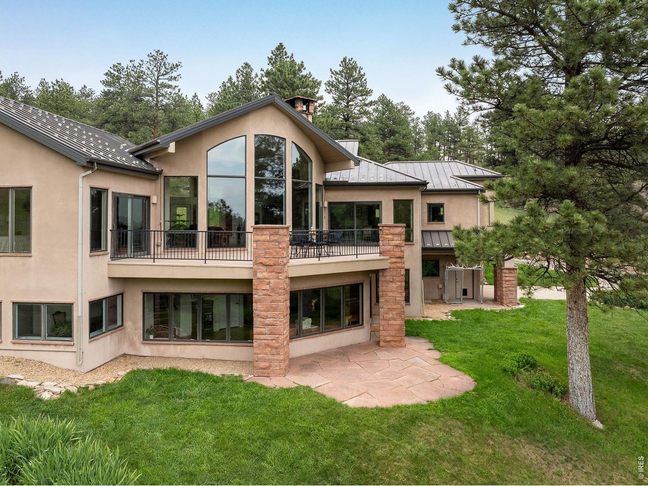Two-story stucco home with a balcony, stone pillars, and a patio among mature pines.