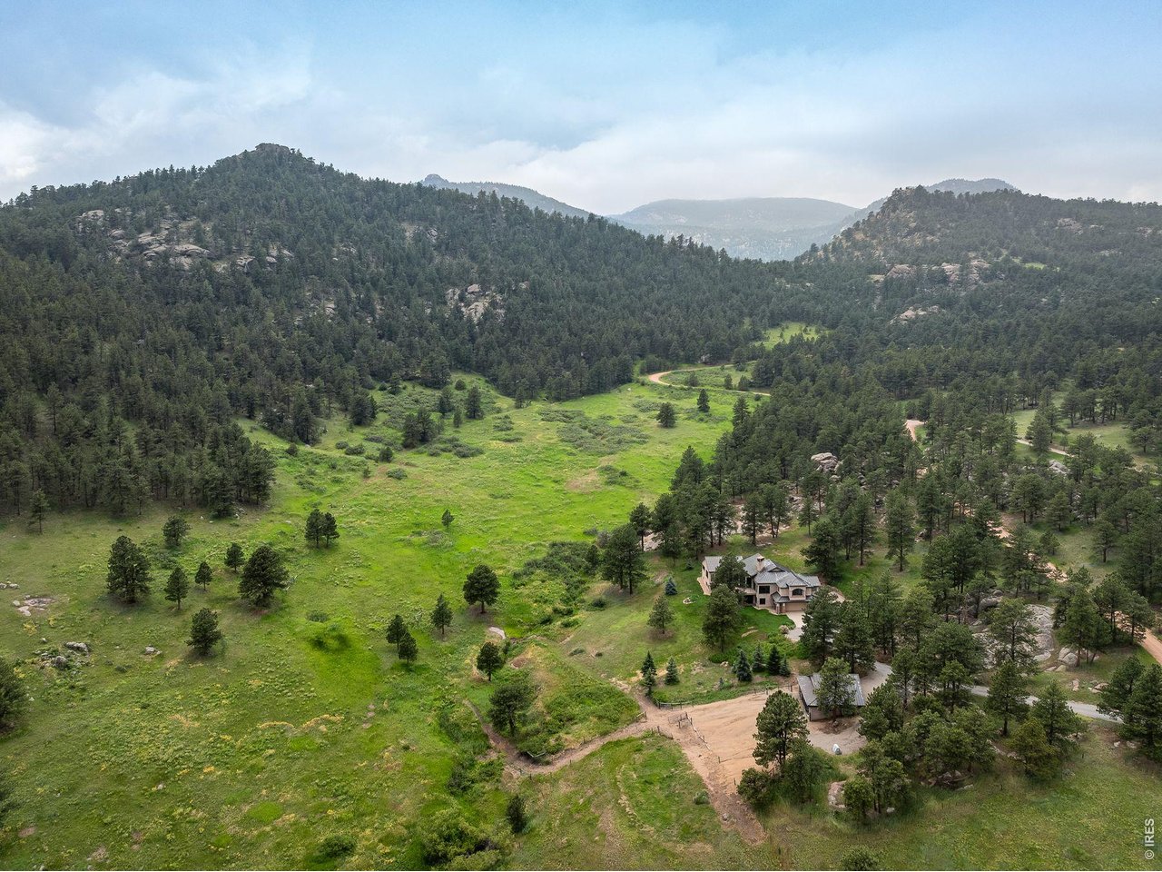 Aerial view of a home in a green mountain meadow surrounded by pine-covered hills.