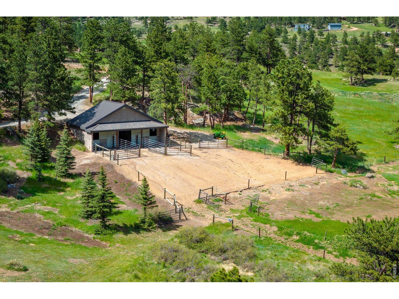 Aerial view of a fenced sandy paddock and metal-roofed stable among pine trees.