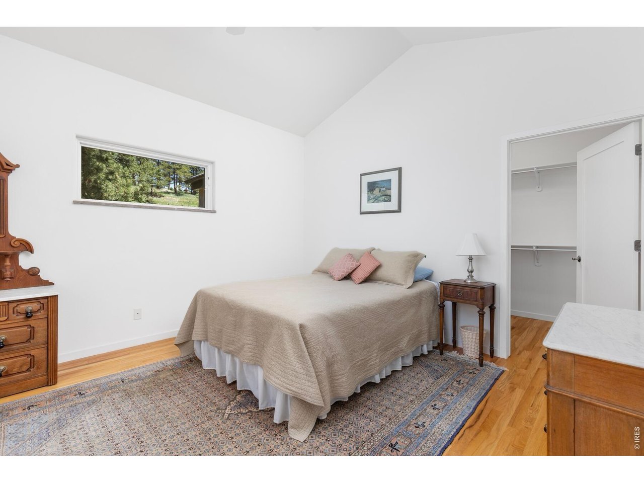 Bedroom with a beige-covered bed, patterned rug, hardwood floors, and a window overlooking trees.