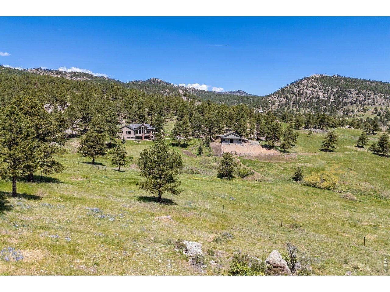 Aerial view of a home and outbuilding on a grassy property amid pine-covered mountain foothills.