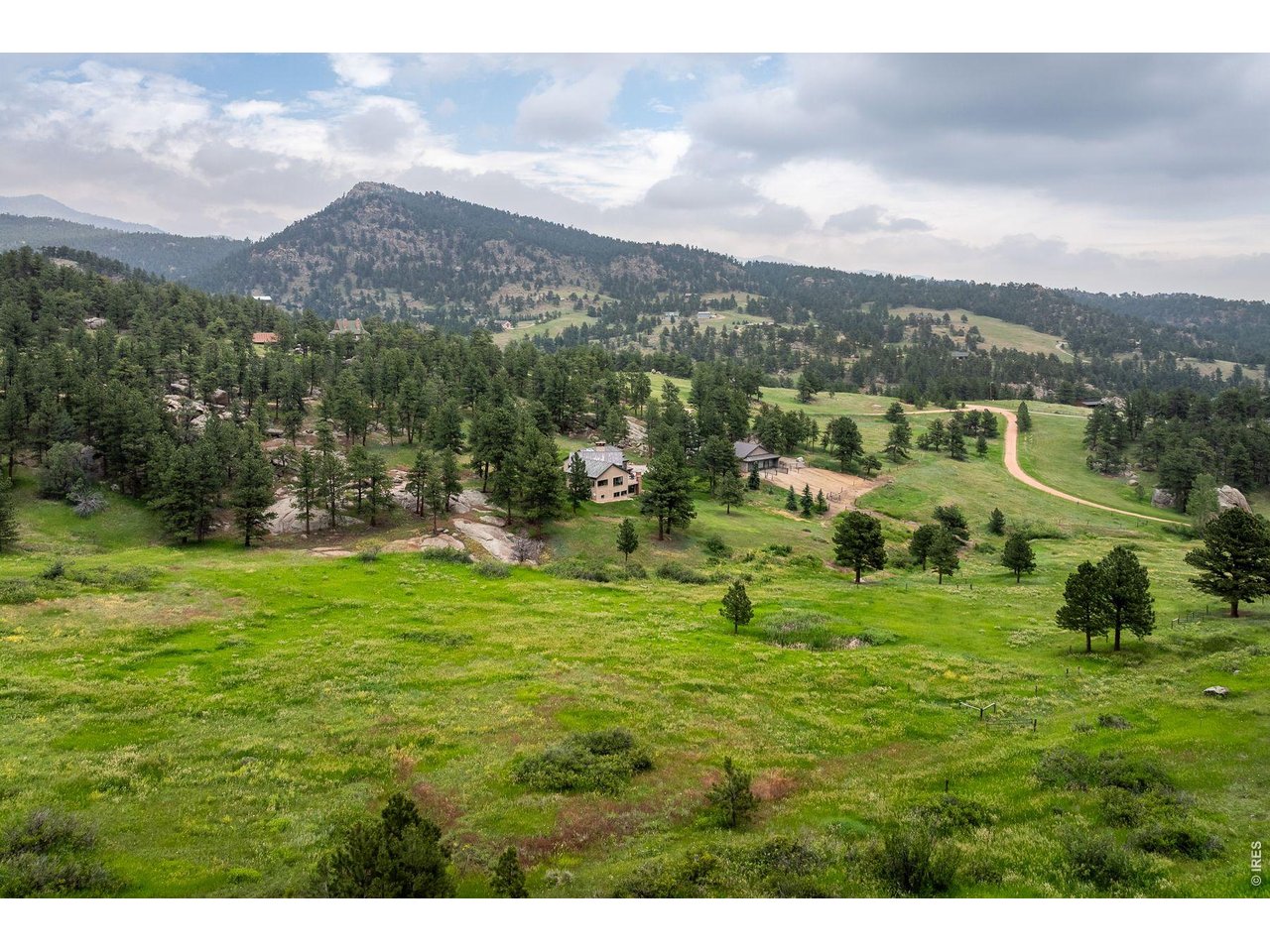 Aerial view of a home amid green meadows and pine-covered hills, with mountains beyond.