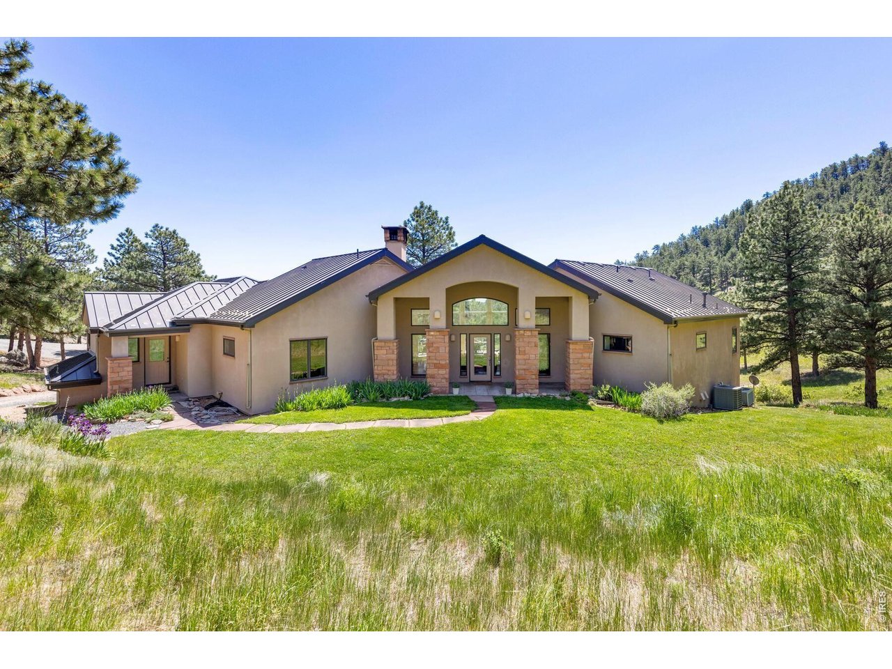 Tan home with a dark metal roof and stone entry columns in a pine-covered hillside setting.