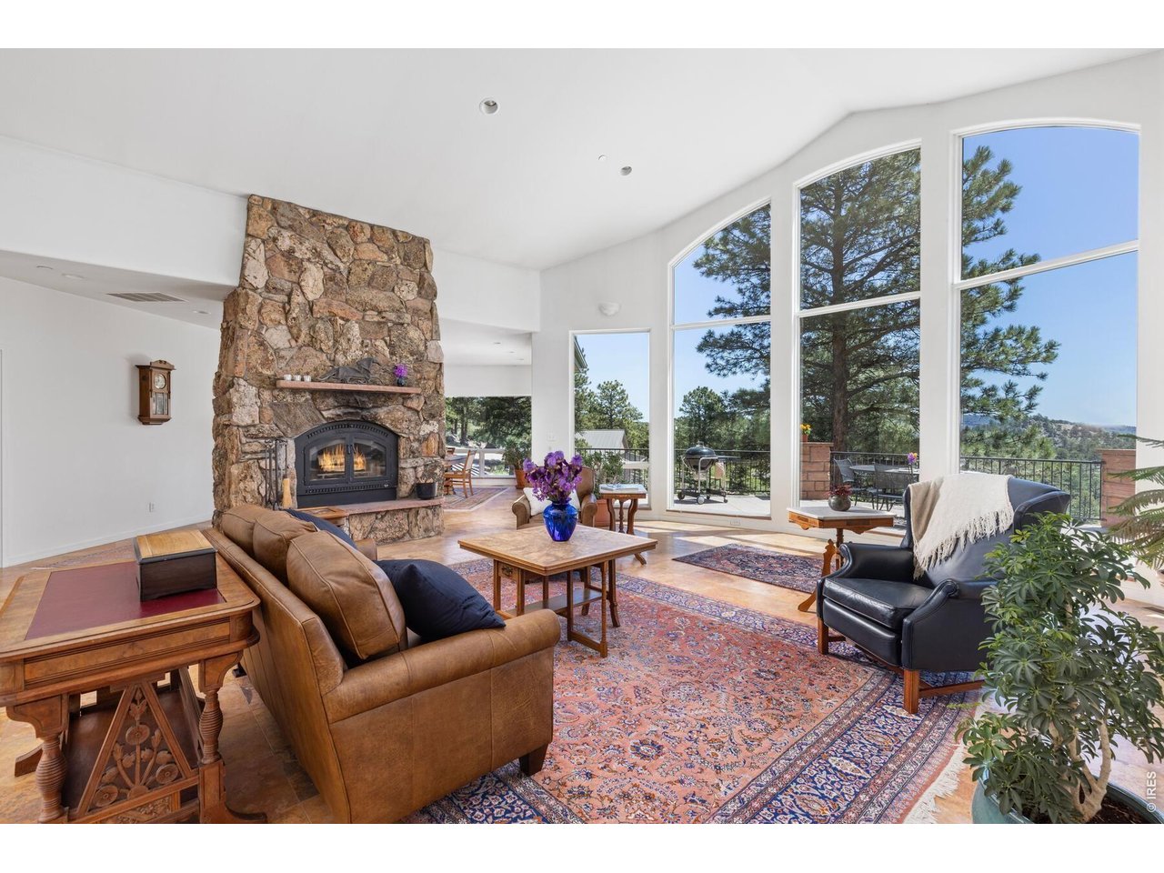 Living room with a stone fireplace, tall arched windows, and views of pine trees.