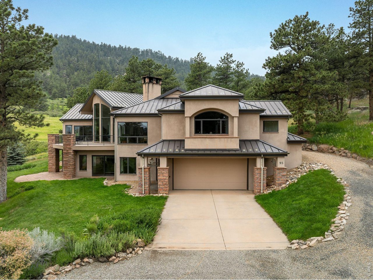 Stucco mountain home with metal roofs, stone columns, driveway, and forested hillside.