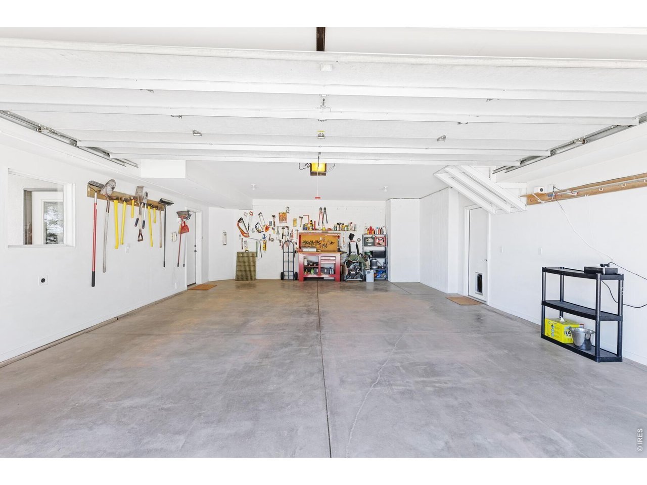 Spacious garage with a concrete floor, raised overhead door, wall-mounted tools, and a workbench.