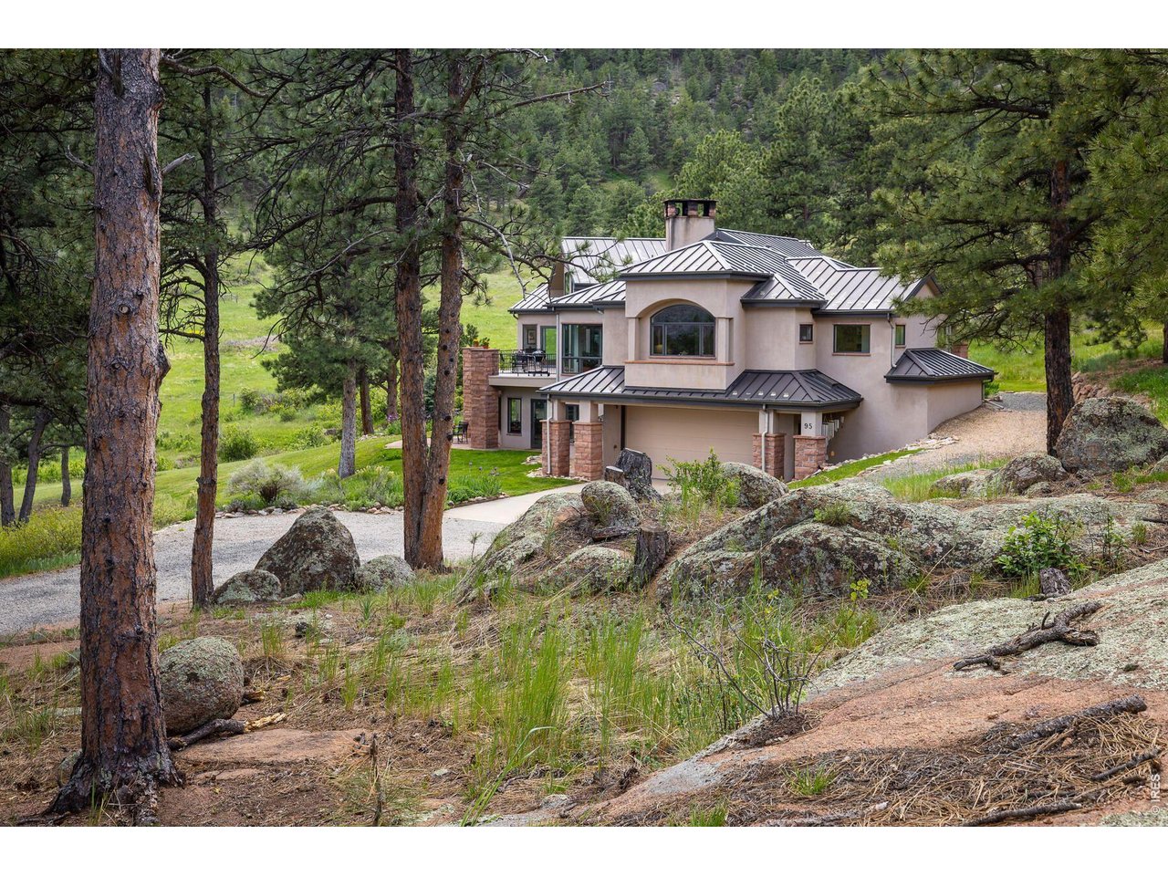 Stucco home with a metal roof among pines and boulders, with a gravel driveway.