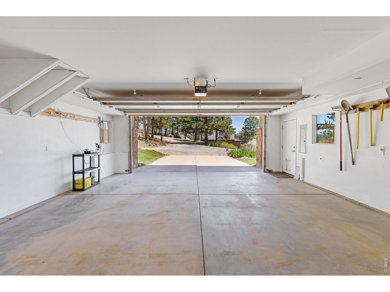 Open two-car garage with a concrete floor, hanging garden tools, and a tree-lined driveway beyond.