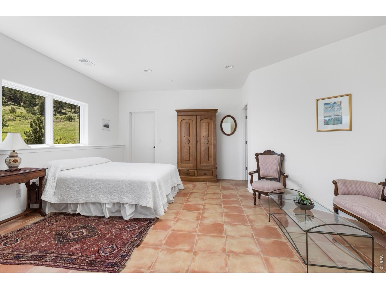 Furnished bedroom with terracotta tile floors, a wooden armoire, and windows overlooking a green hillside.