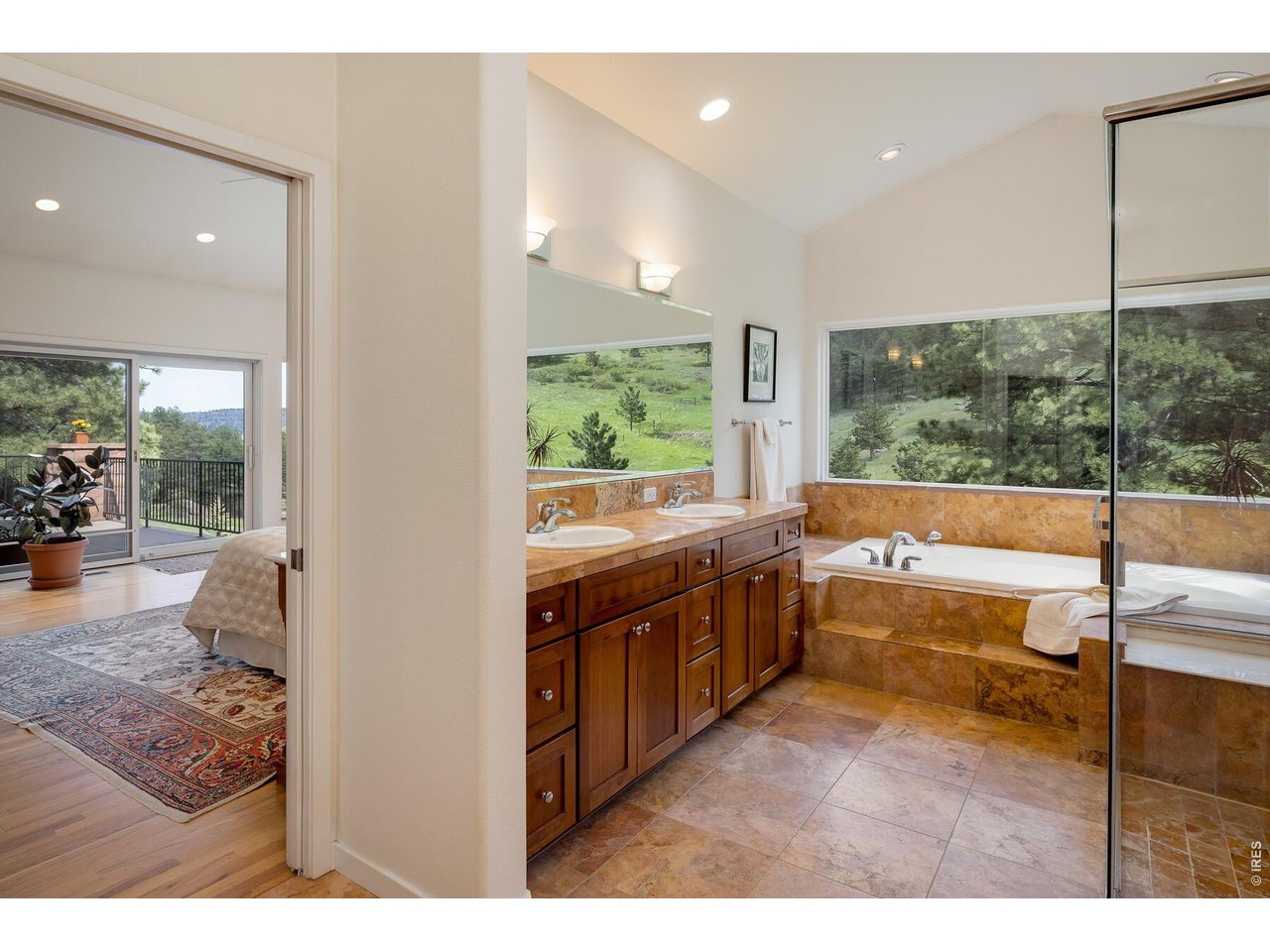 Bathroom with a double vanity and soaking tub beside large windows overlooking a wooded hillside.