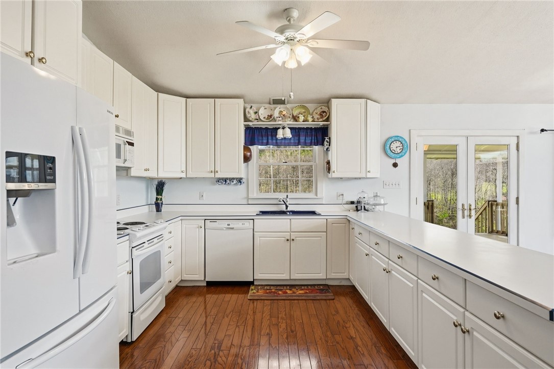 White kitchen with L-shaped counters, wood flooring, and glass doors opening to a wooded view.
