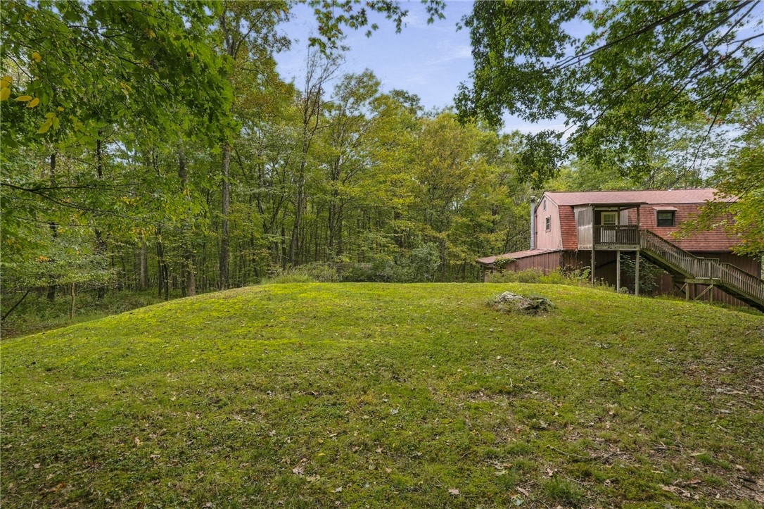 House with an elevated wooden deck and staircase beside a grassy yard bordered by mature trees.