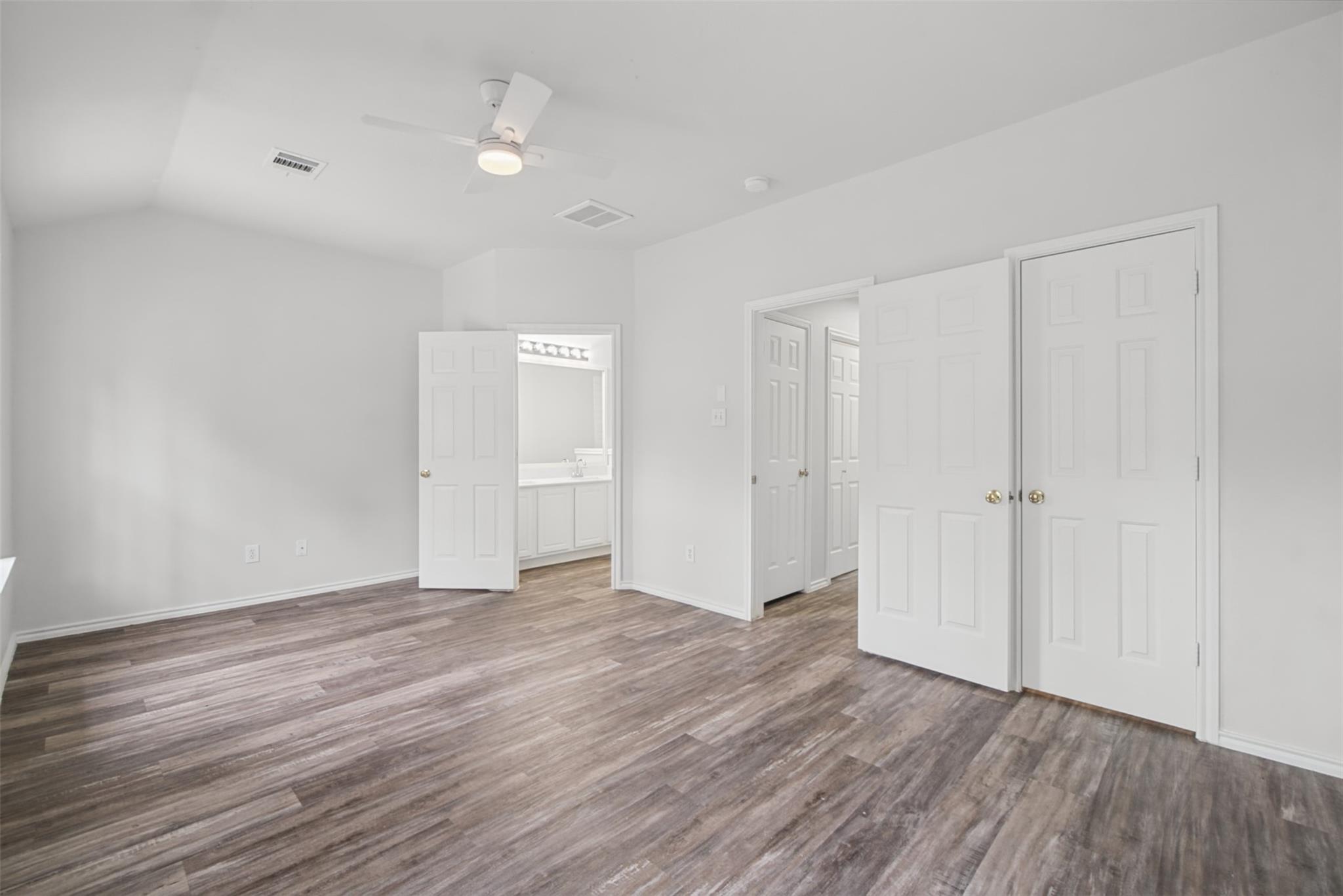 Empty bedroom with plank flooring, ceiling fan, and open doors to a bathroom and hallway.