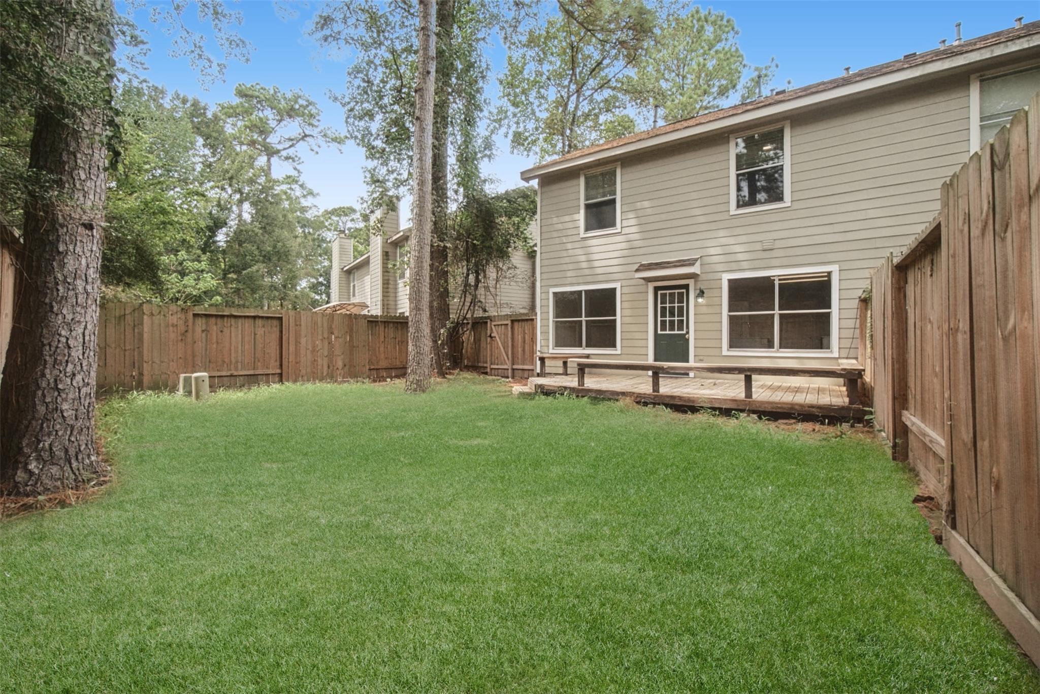 Two-story beige house behind a fenced lawn, with a small wooden deck and mature trees.