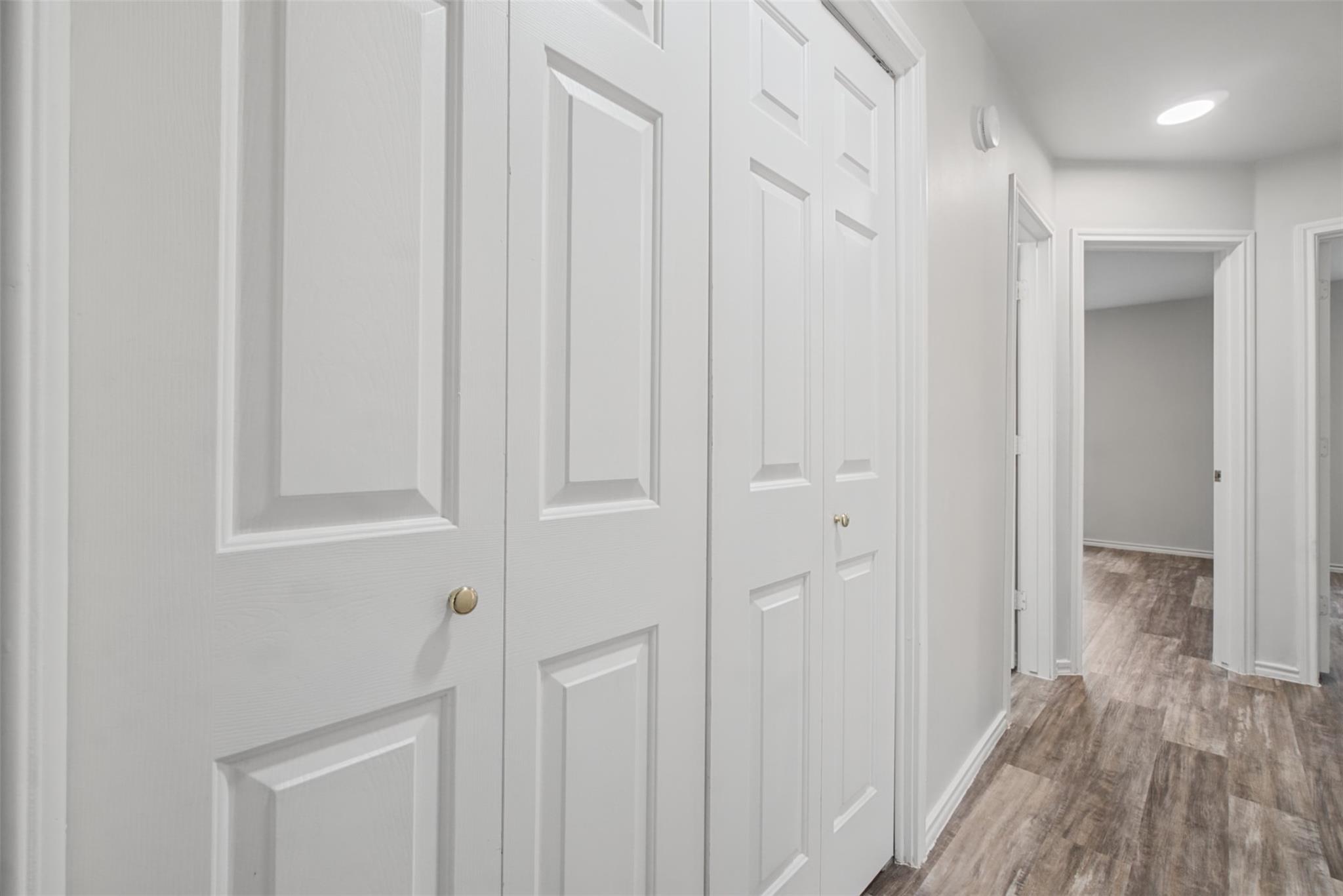 Hallway with white paneled closet doors and wood-look flooring leading to an open doorway.