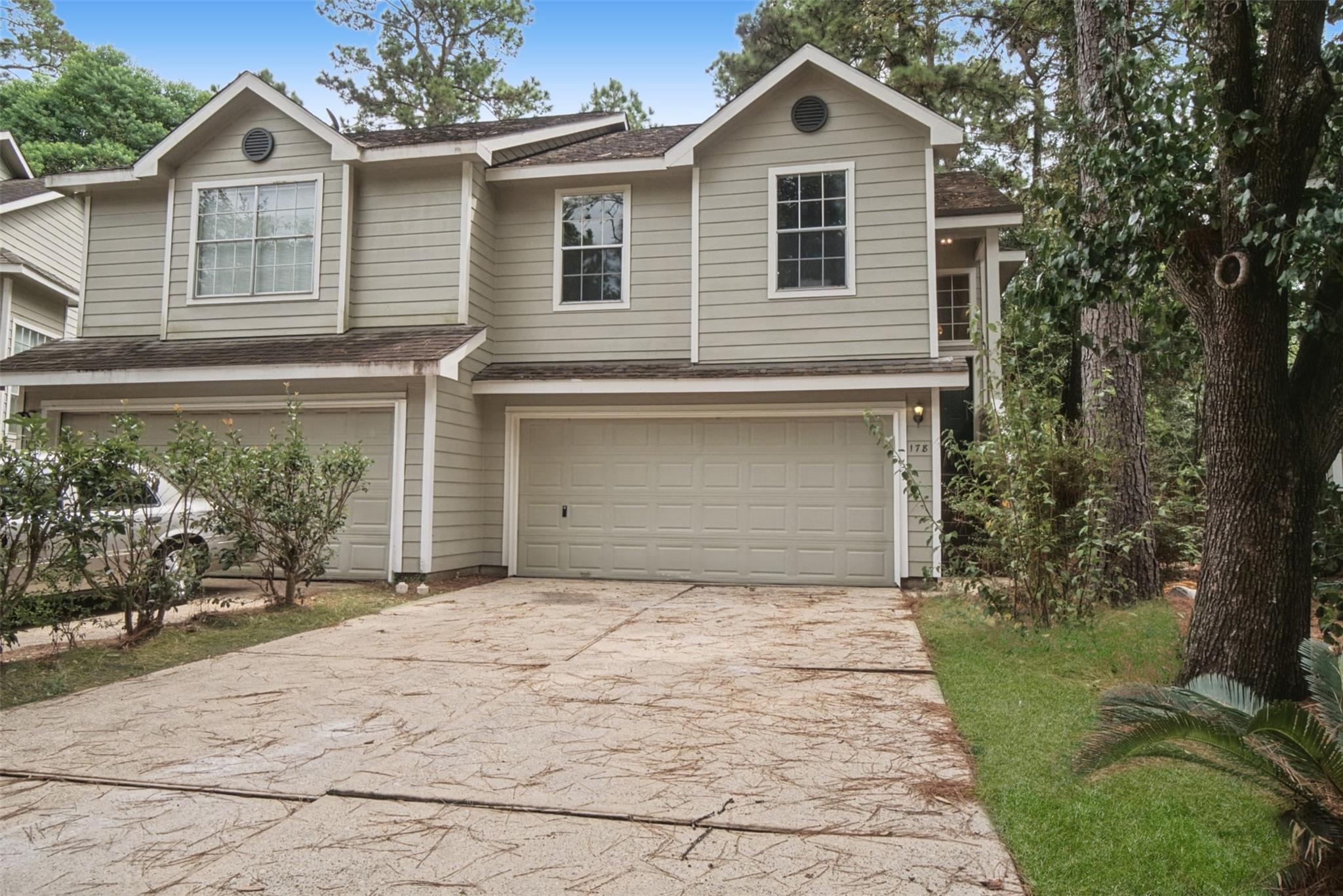 Two-story beige house with two garage doors, a broad driveway, and mature trees.