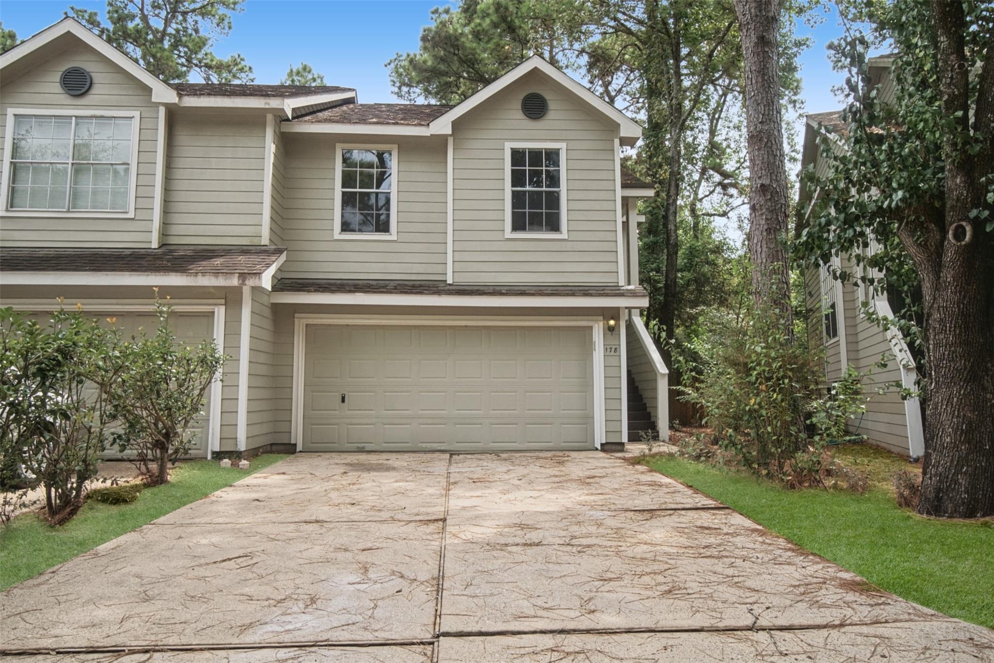 Two-story beige house with a double garage, broad concrete driveway, and mature trees.