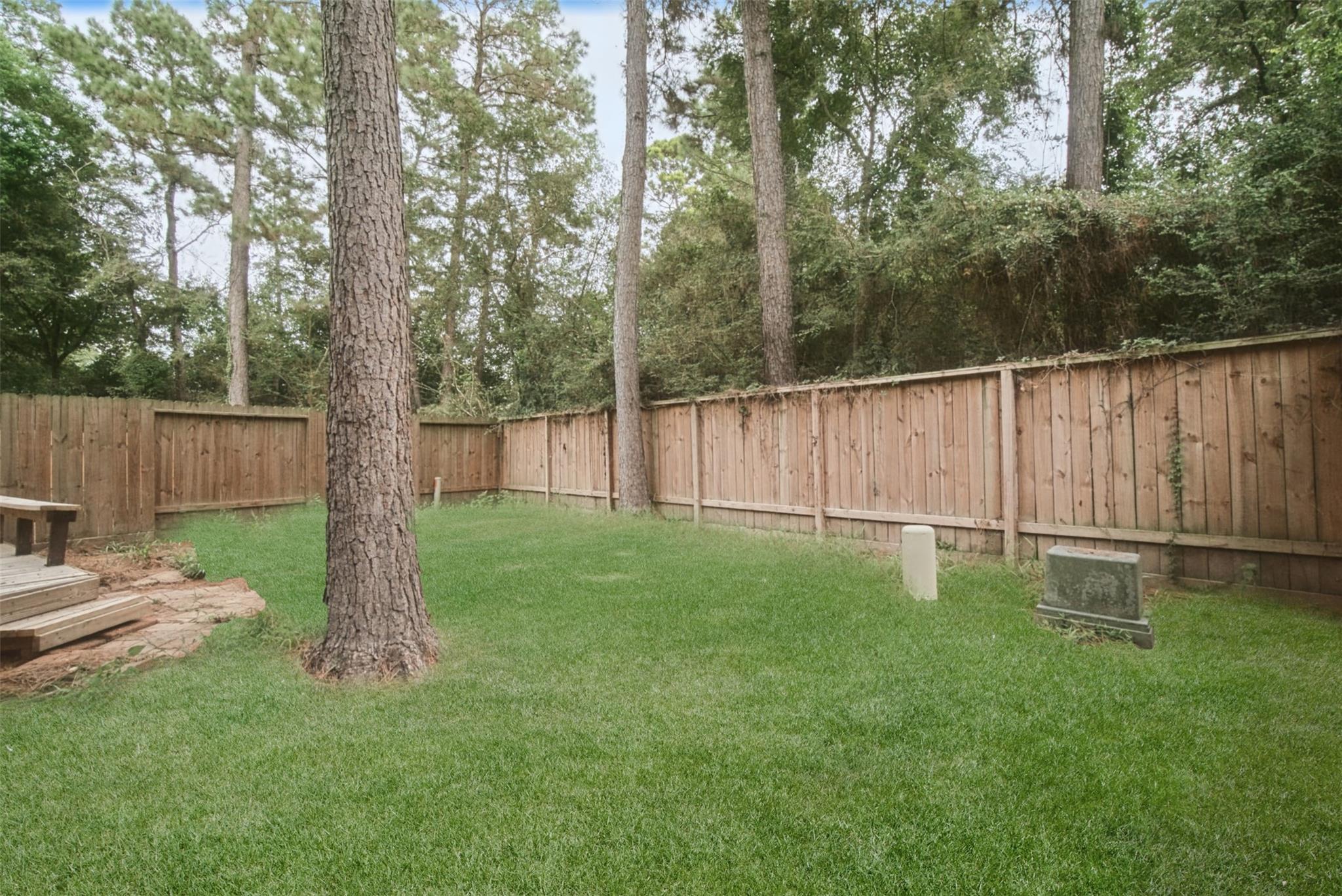 Fenced backyard with a green lawn, mature pine trees, and a small wooden deck.