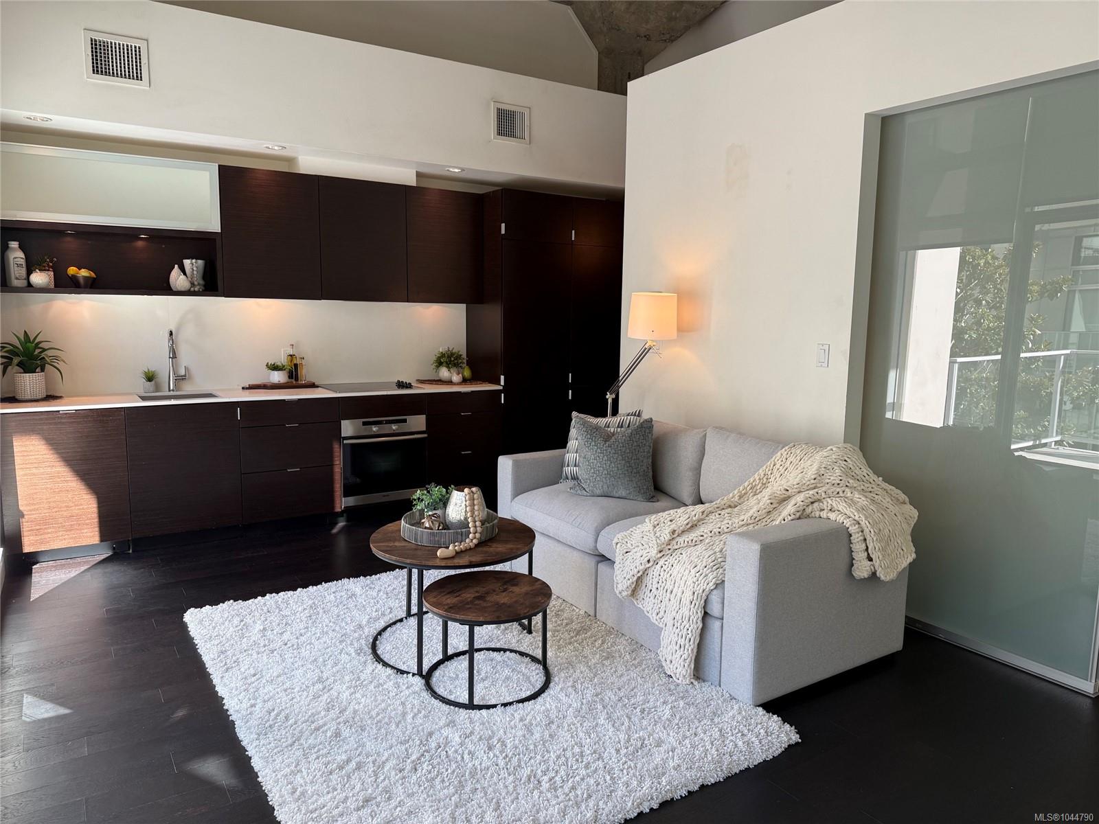 Open-plan condo kitchen and living area with dark cabinetry, a gray sofa, and large windows.
