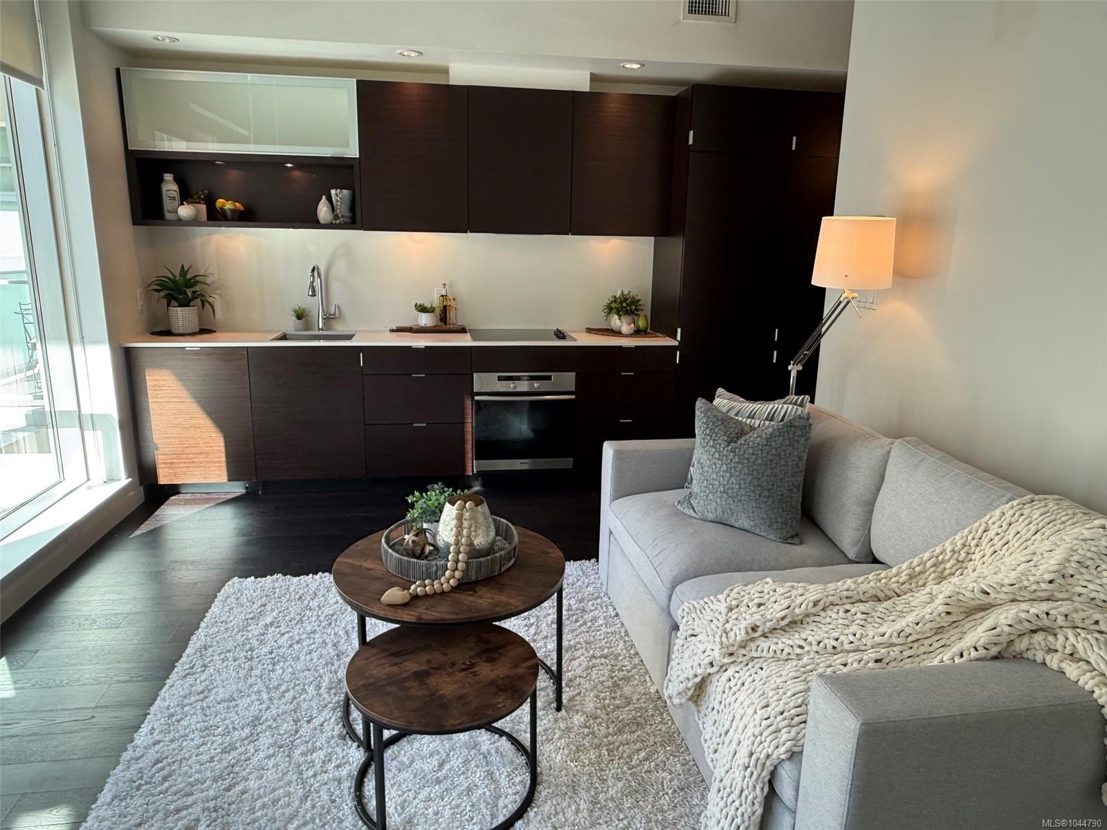 Open-plan condo living room and kitchen with dark cabinetry, a gray sofa, and large windows.