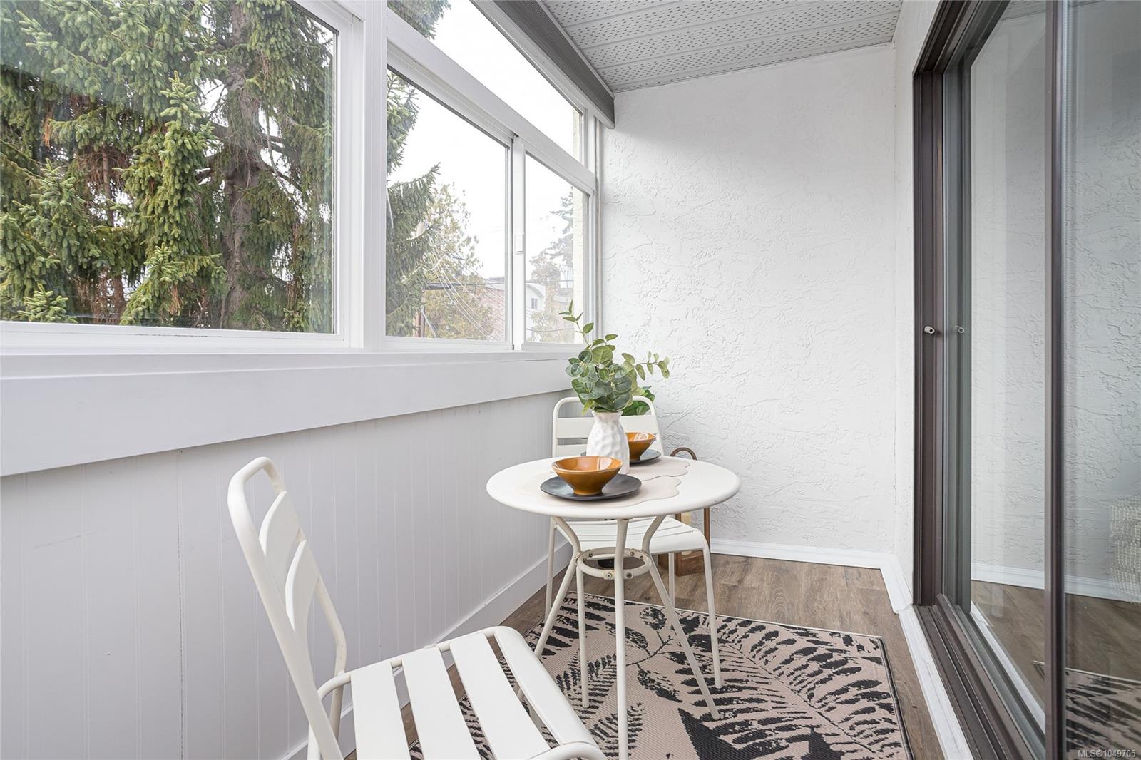 Enclosed balcony with white seating, a round table, and windows overlooking evergreen trees.