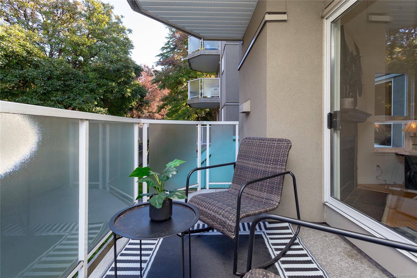 Apartment balcony with frosted glass railings, woven chair, potted plant, and leafy trees beyond.