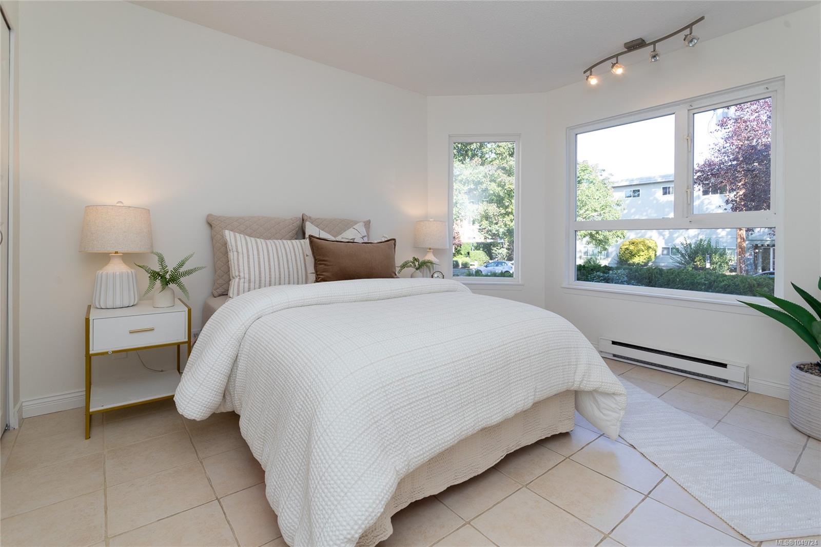 Staged bedroom with a large white bed, tiled floor, and windows overlooking trees.