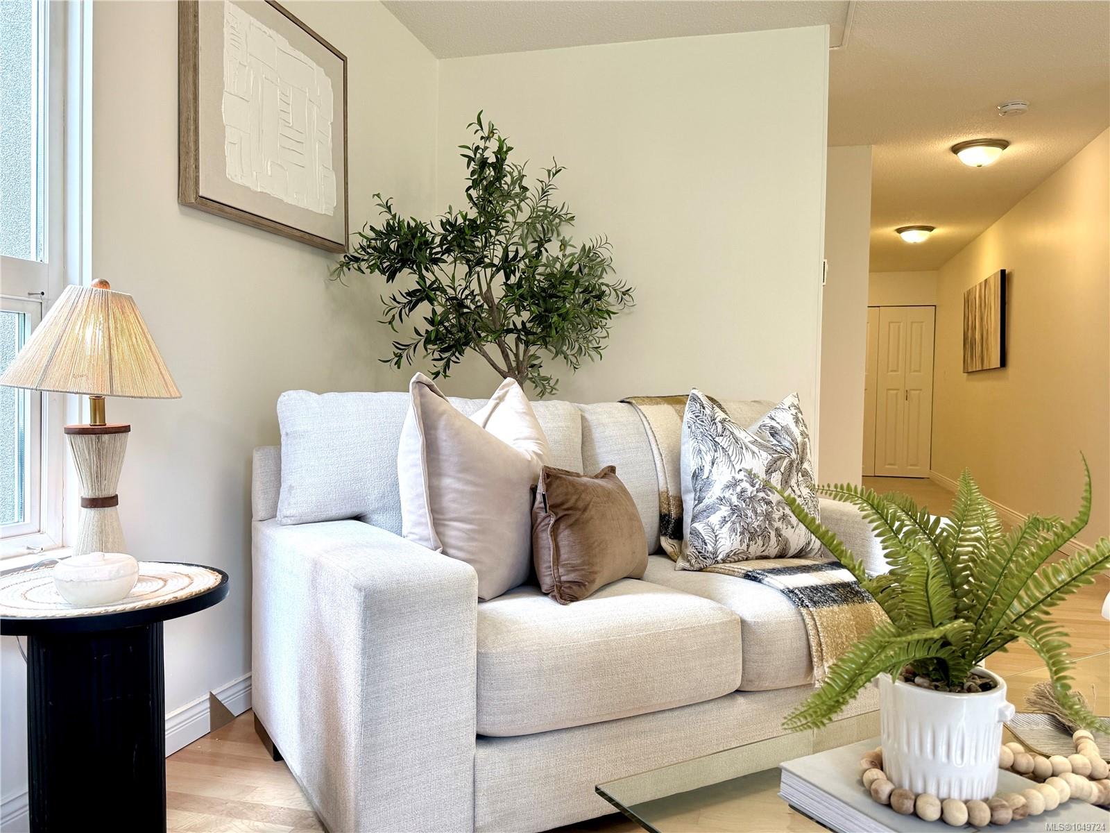 Staged living room with a light sofa, leafy plants, and a hallway in the background.