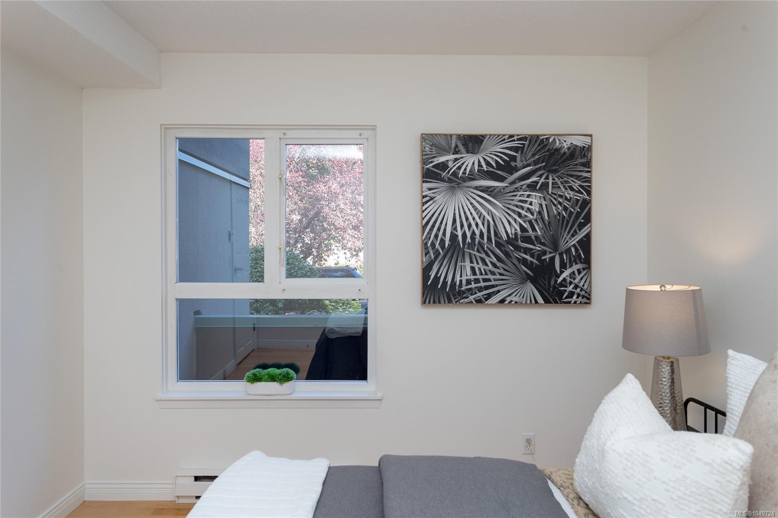 Staged bedroom with a gray bed, botanical wall art, and a large window overlooking leafy trees.