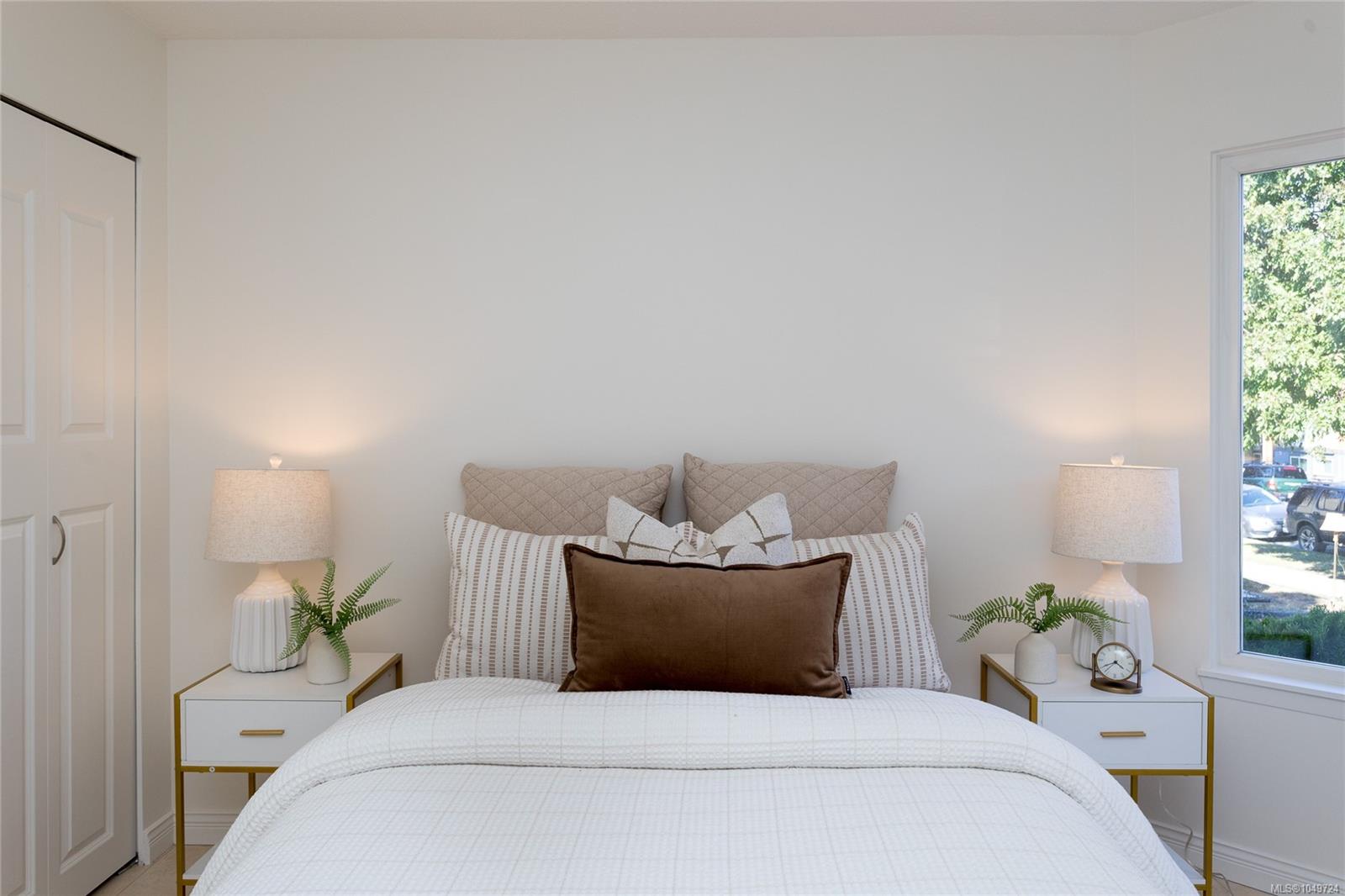 Staged bedroom with a white bed, layered neutral pillows, matching bedside tables, and a large window.