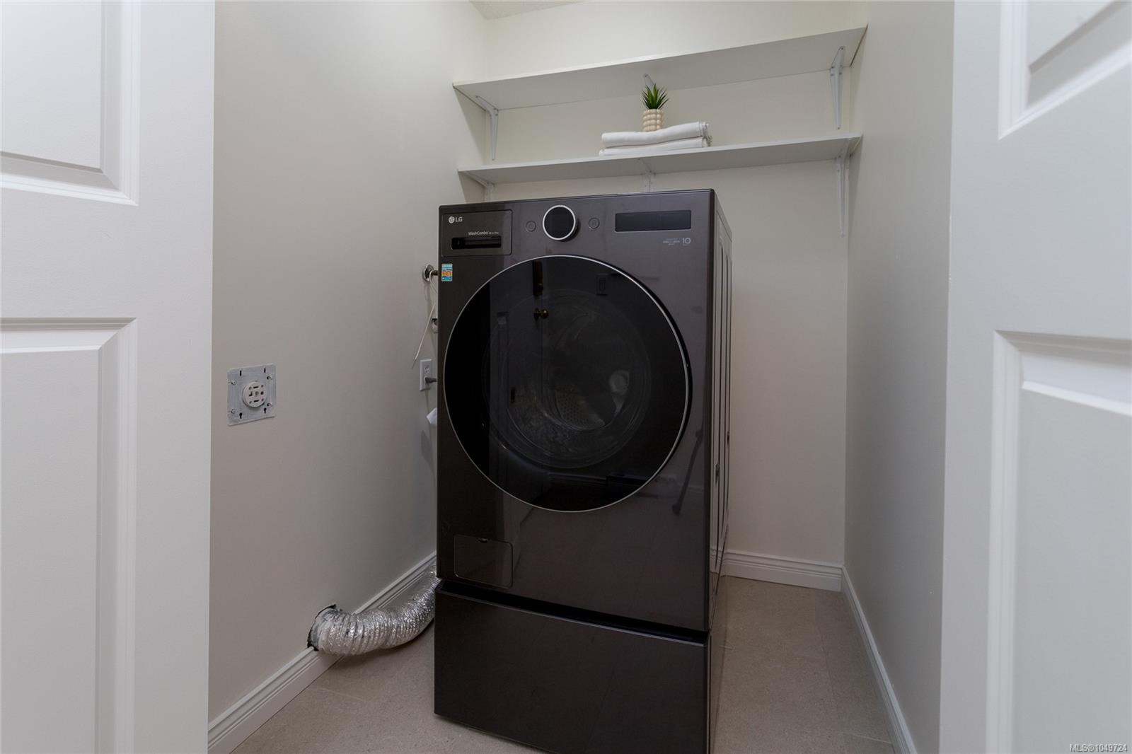 Laundry closet with a dark front-loading washer-dryer combo beneath shelves holding towels and a small plant.