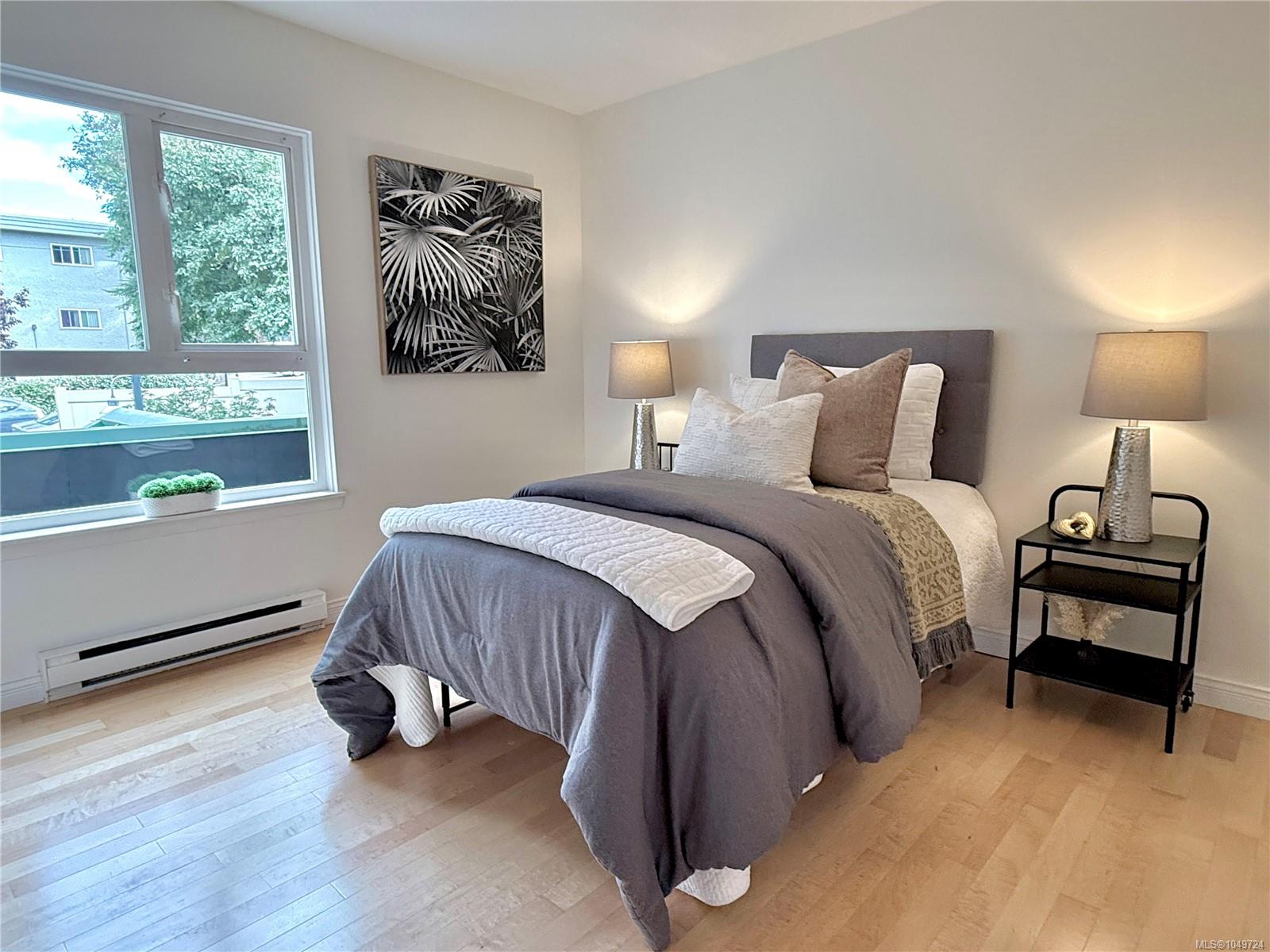Staged bedroom with gray bedding, matching bedside lamps, and a large window overlooking trees.