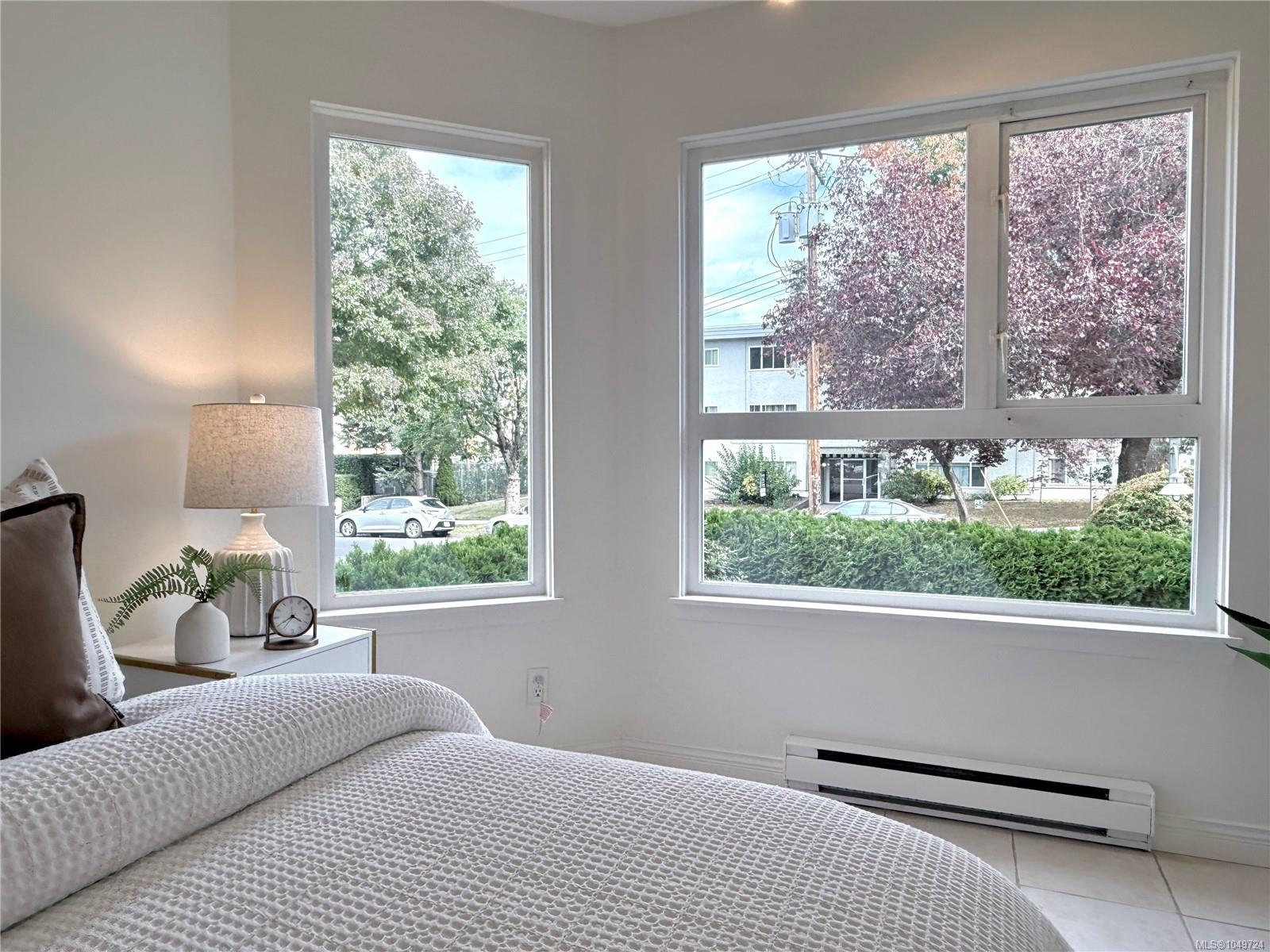 Furnished bedroom with a white textured bedspread and large windows overlooking leafy trees.