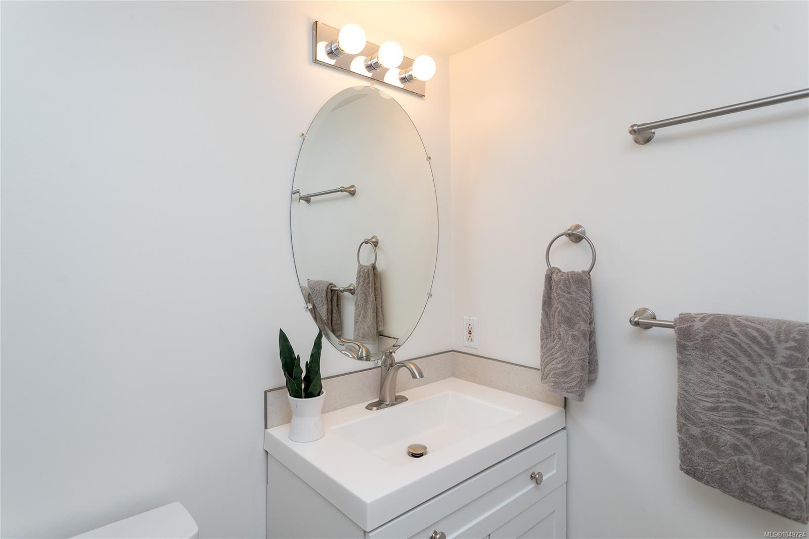 Bathroom vanity with an oval mirror, three-bulb light, chrome fixtures, gray towels, and a potted plant.