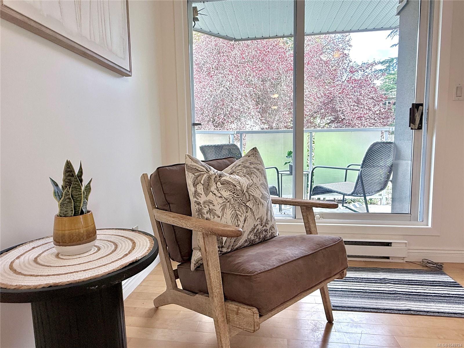 Living room with a wooden armchair beside large windows overlooking a furnished balcony and leafy trees.