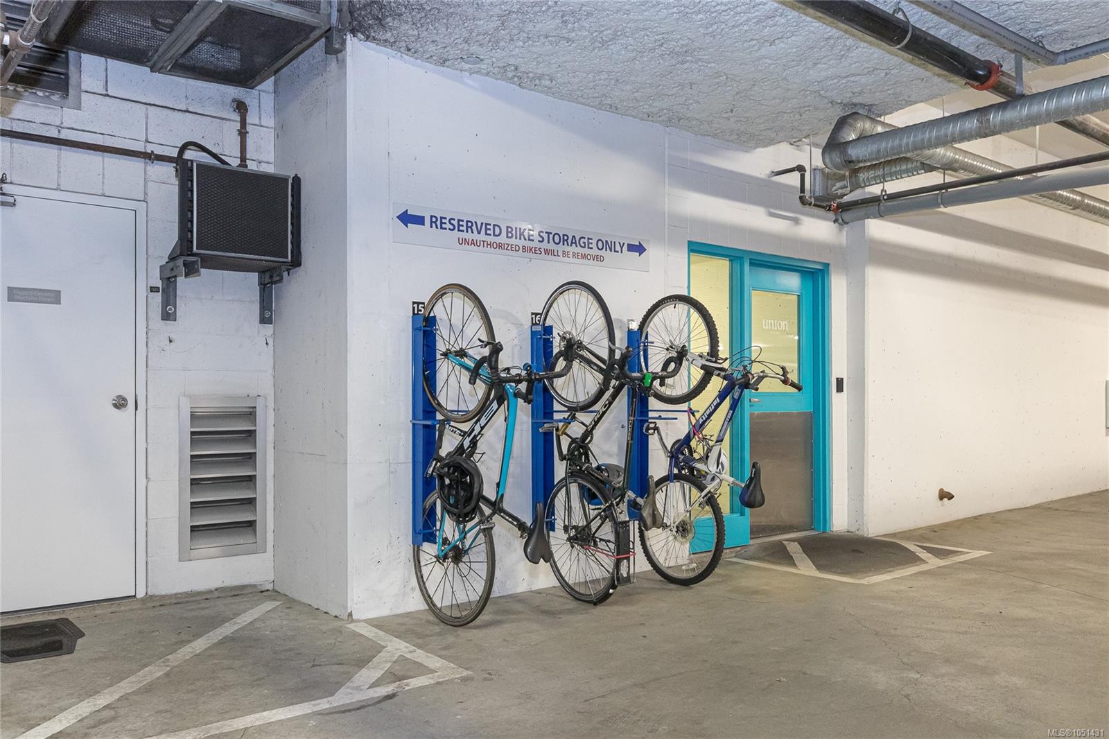 Shared bike storage area with bicycles in blue racks beside a turquoise-framed door.