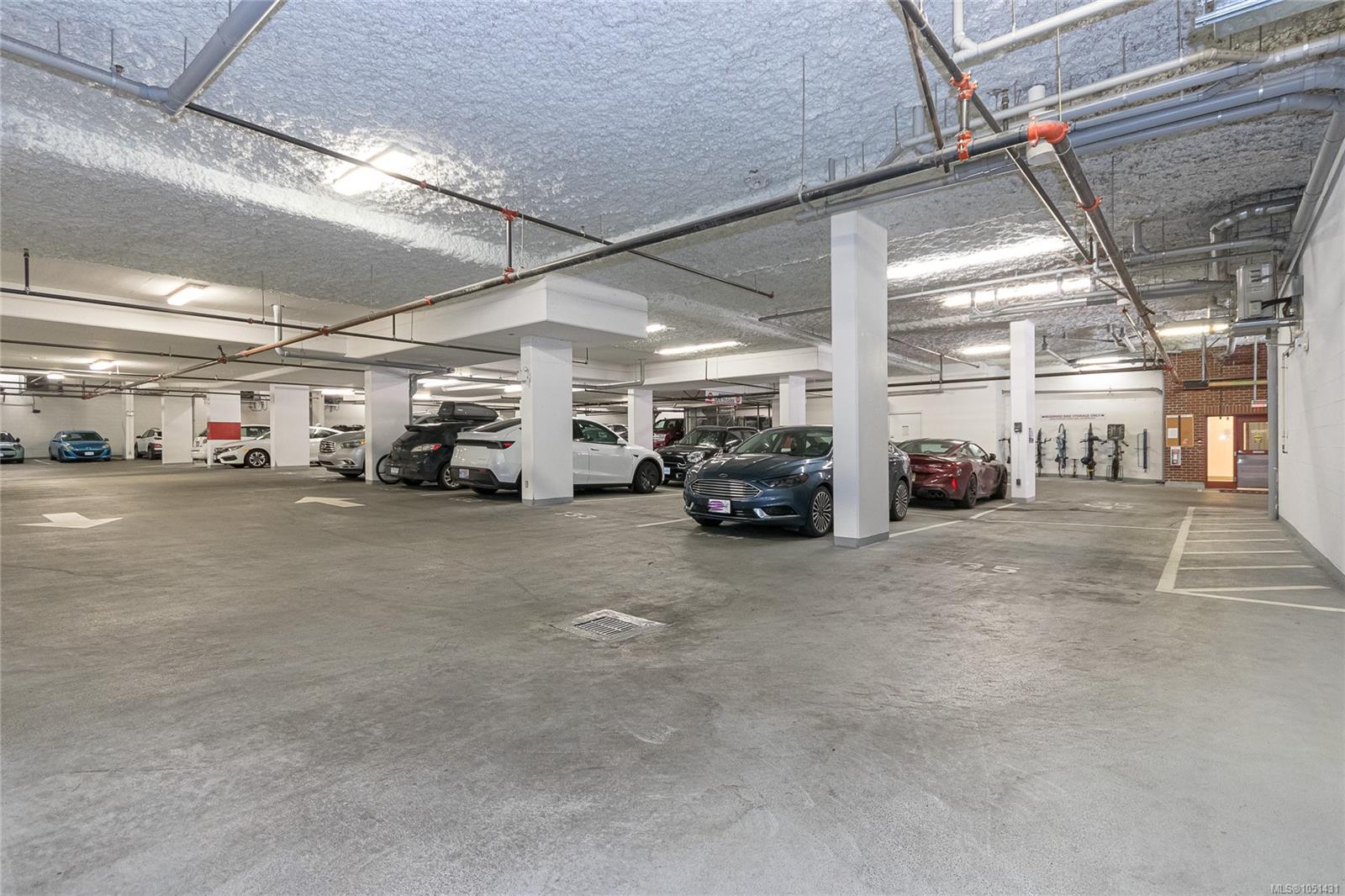 Underground parking garage with concrete pillars, exposed pipes, and several parked cars.