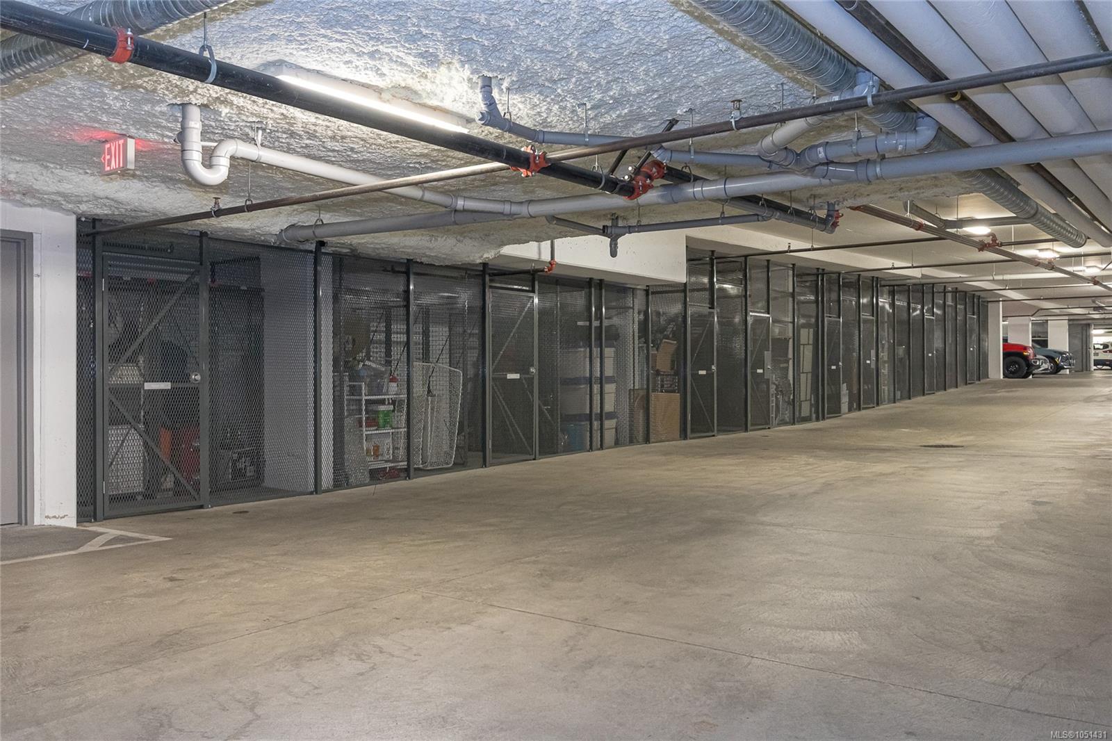 Underground parking garage with mesh storage lockers, exposed ceiling pipes, and a concrete driving aisle.