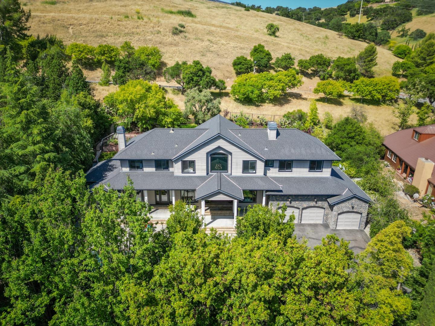 Aerial view of a large gray two-story house with stone-fronted garages, surrounded by trees and dry hills.