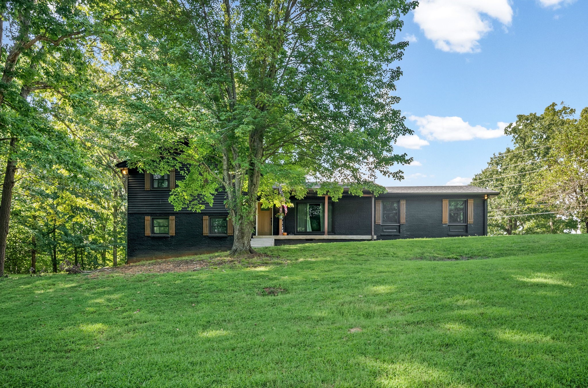 Dark-sided house with a long porch beneath mature trees, overlooking a broad sloping lawn.